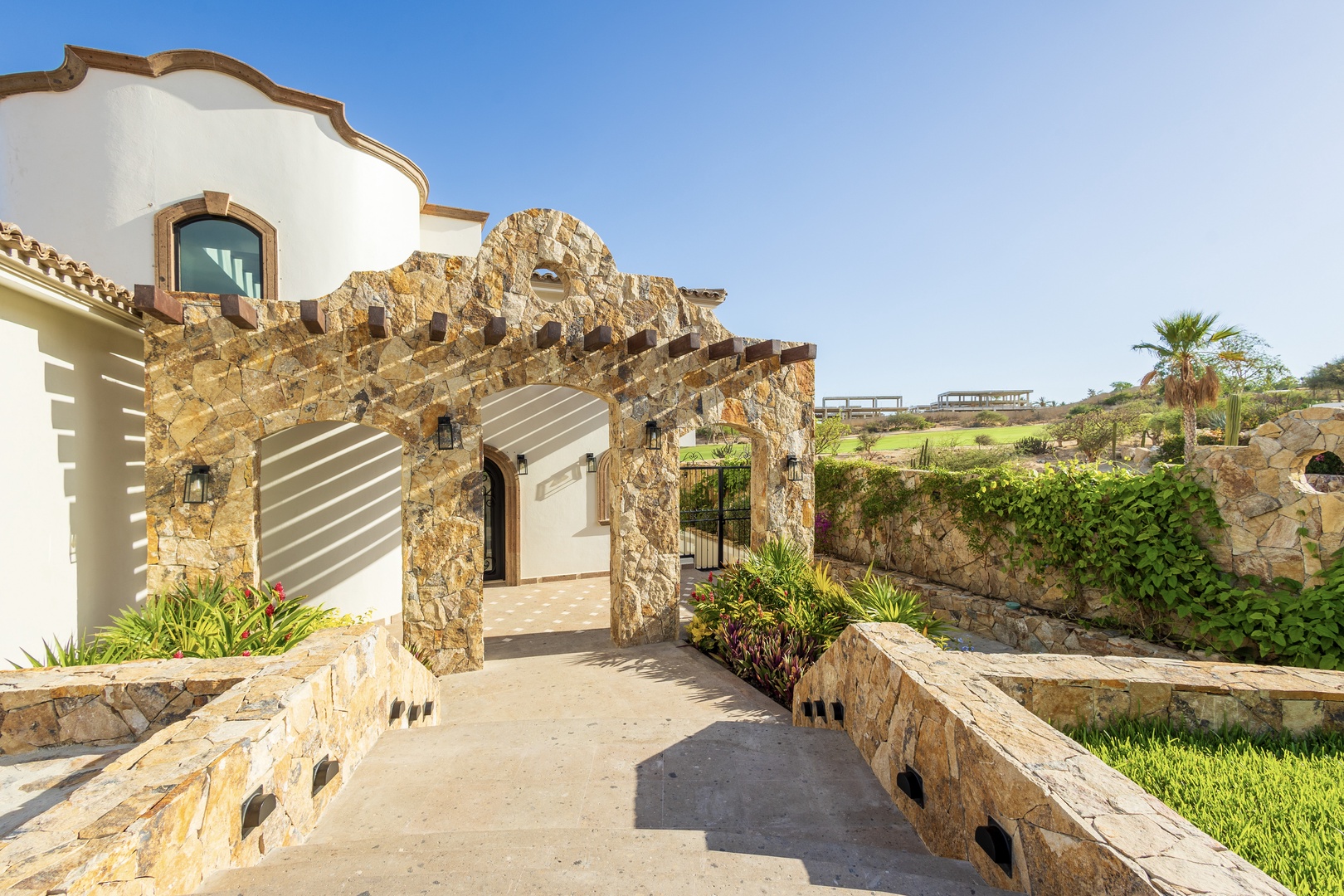 This charming stone archway frames the entrance to a beautifully designed villa with Spanish-colonial accents, surrounded by lush desert landscaping and open views of the countryside.