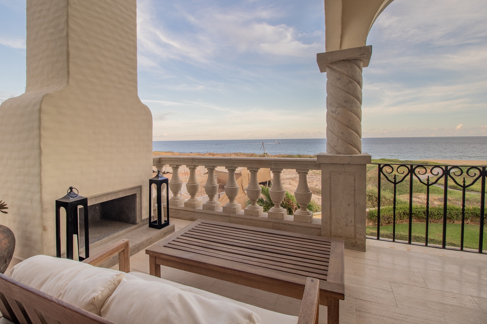 Elegant beachfront terrace with Mediterranean architecture overlooking calm ocean waters. A peaceful outdoor lounge space with decorative columns, fireplace, and direct views of the waterfront and surrounding gardens.