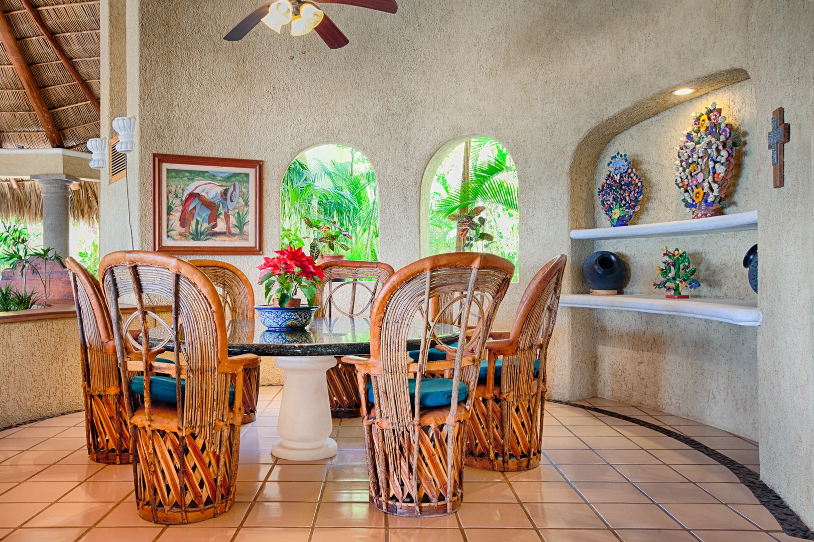 Enjoy tropical elegance in your dining space with handcrafted wicker chairs, vibrant artwork, and lush garden views through arched windows.