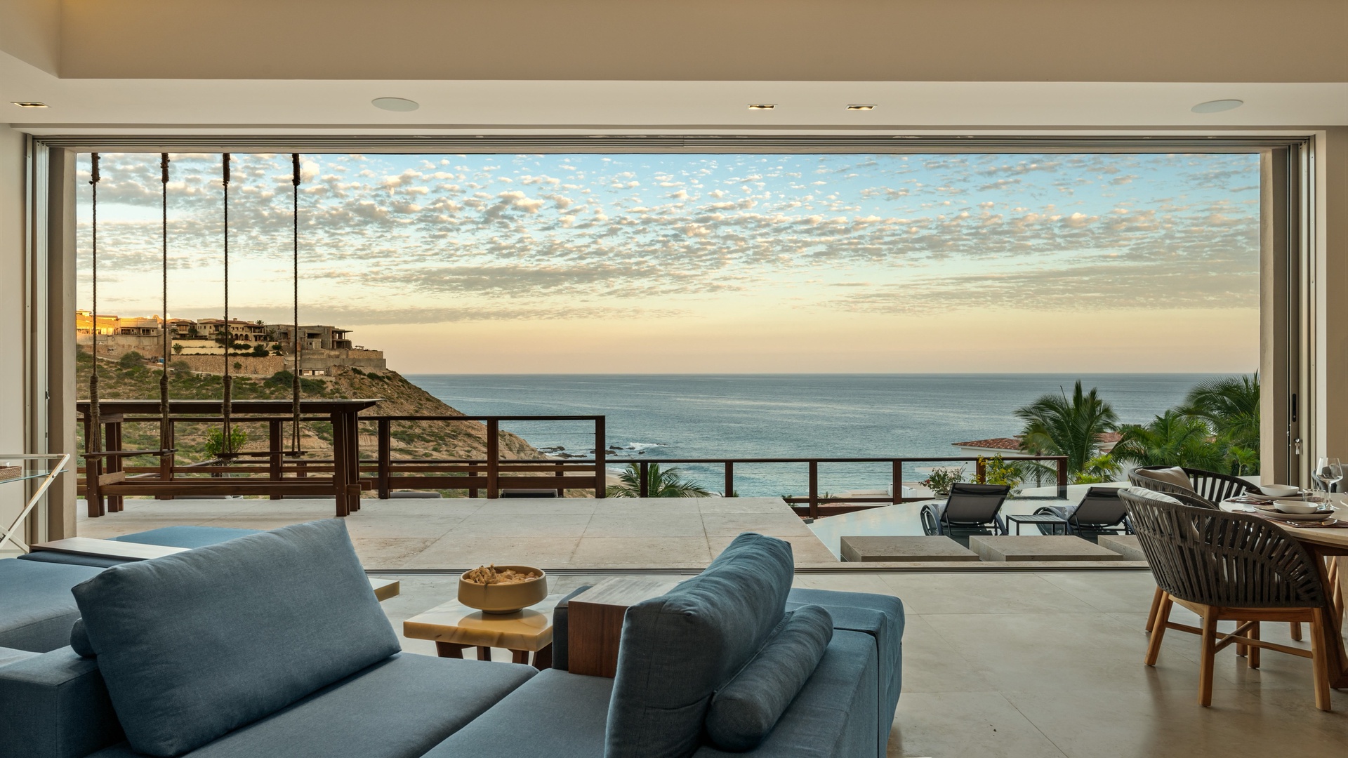 A stunning coastal property perched on a hillside, framed by palm trees and offering an expansive terrace overlooking the ocean at sunset.