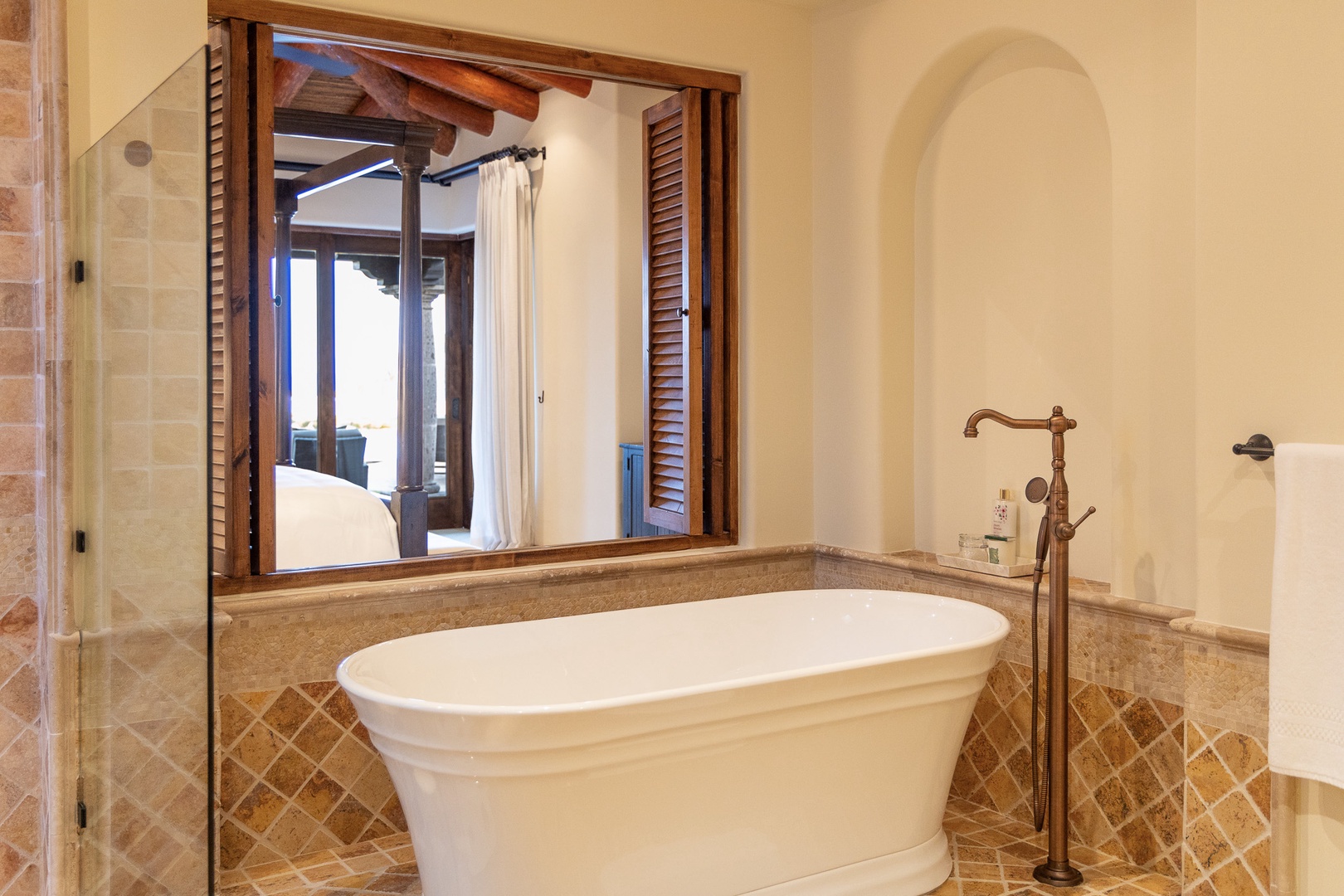 Sink into your elegant soaking tub, surrounded by warm stone and arched alcoves that echo Mediterranean charm, with a window framing peaceful views of the bedroom beyond.