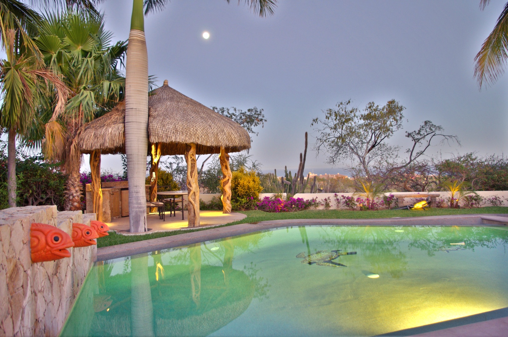 This tranquil backyard oasis features a sparkling pool illuminated at dusk, surrounded by lush tropical landscaping and a charming thatched palapa—a perfect setting for relaxation and evening gatherings.