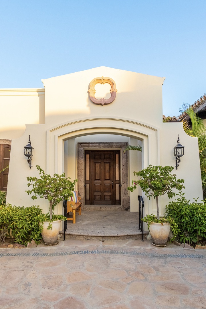 An elegant entrance with white stucco walls, ornate carved detail, and lush greenery frames this property's welcoming doorway.