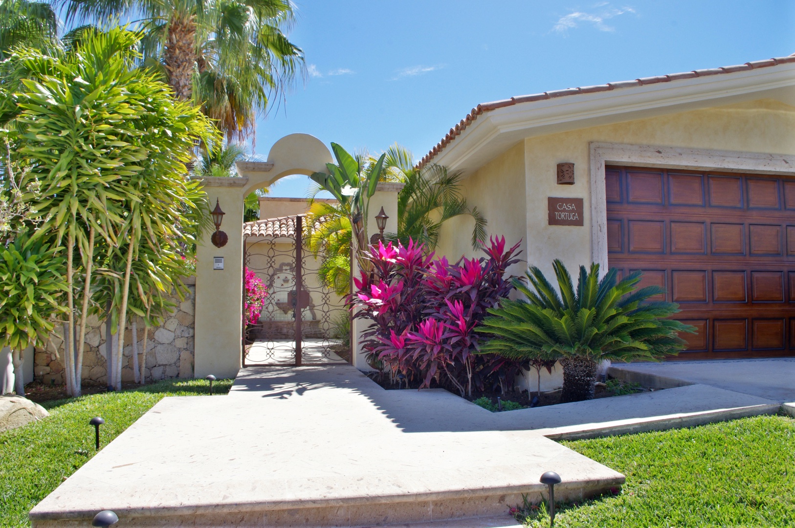 A Mediterranean-style property entrance features lush tropical landscaping and vibrant pink foliage. The archway entrance and stucco facade welcome visitors to this sunny vacation retreat.