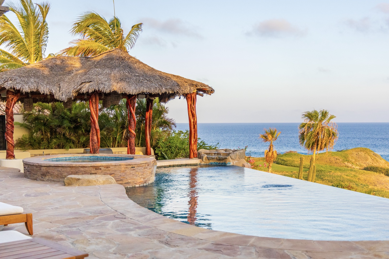 This stunning infinity pool with thatched palapa and hot tub overlooks the sparkling sea, surrounded by swaying palms and coastal beauty—a perfect tropical paradise setting.
