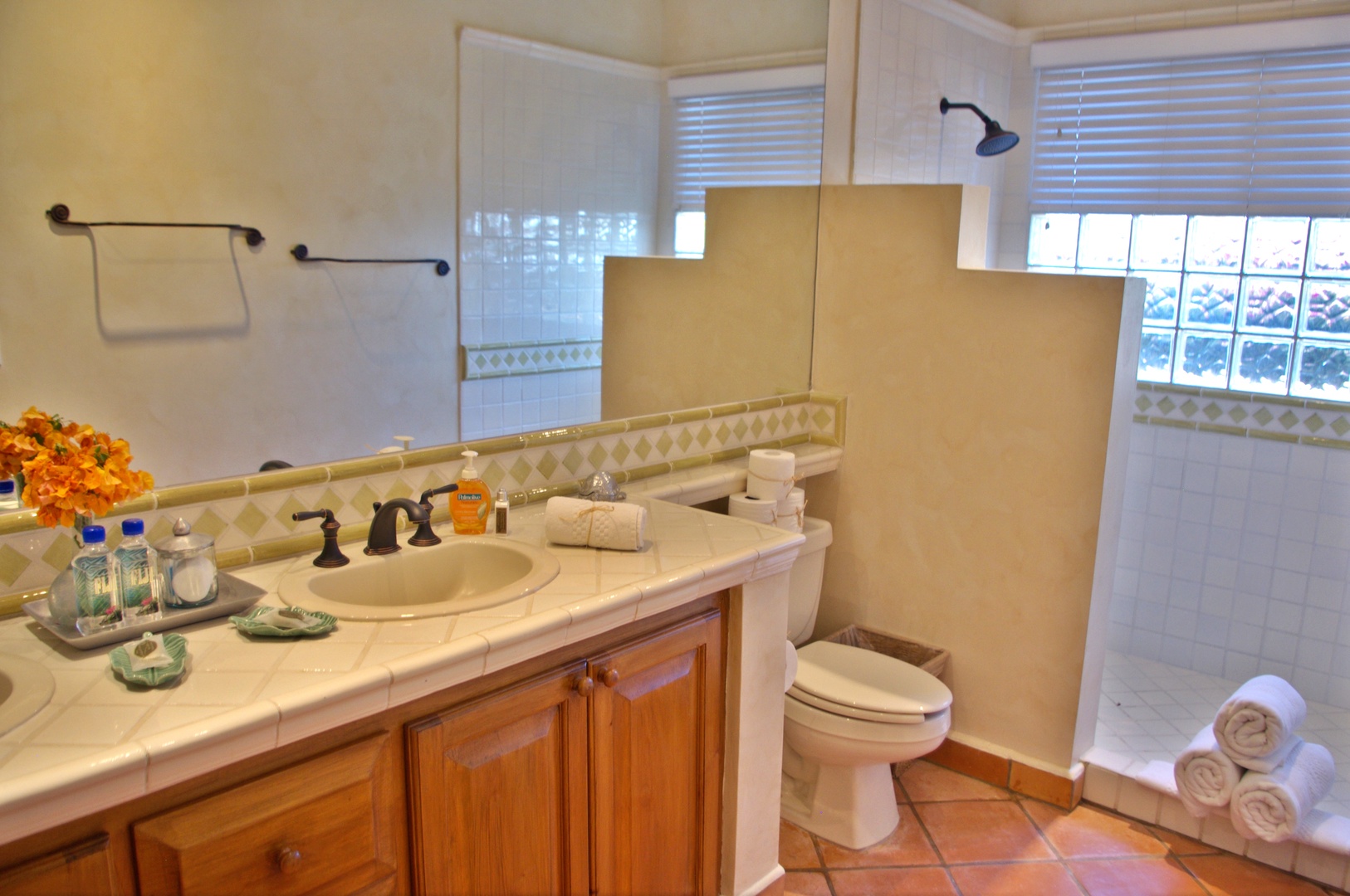 Your private bathroom features a spacious walk-in shower, elegant tiled countertop with fresh amenities, and warm terracotta flooring that adds a cozy, inviting touch.