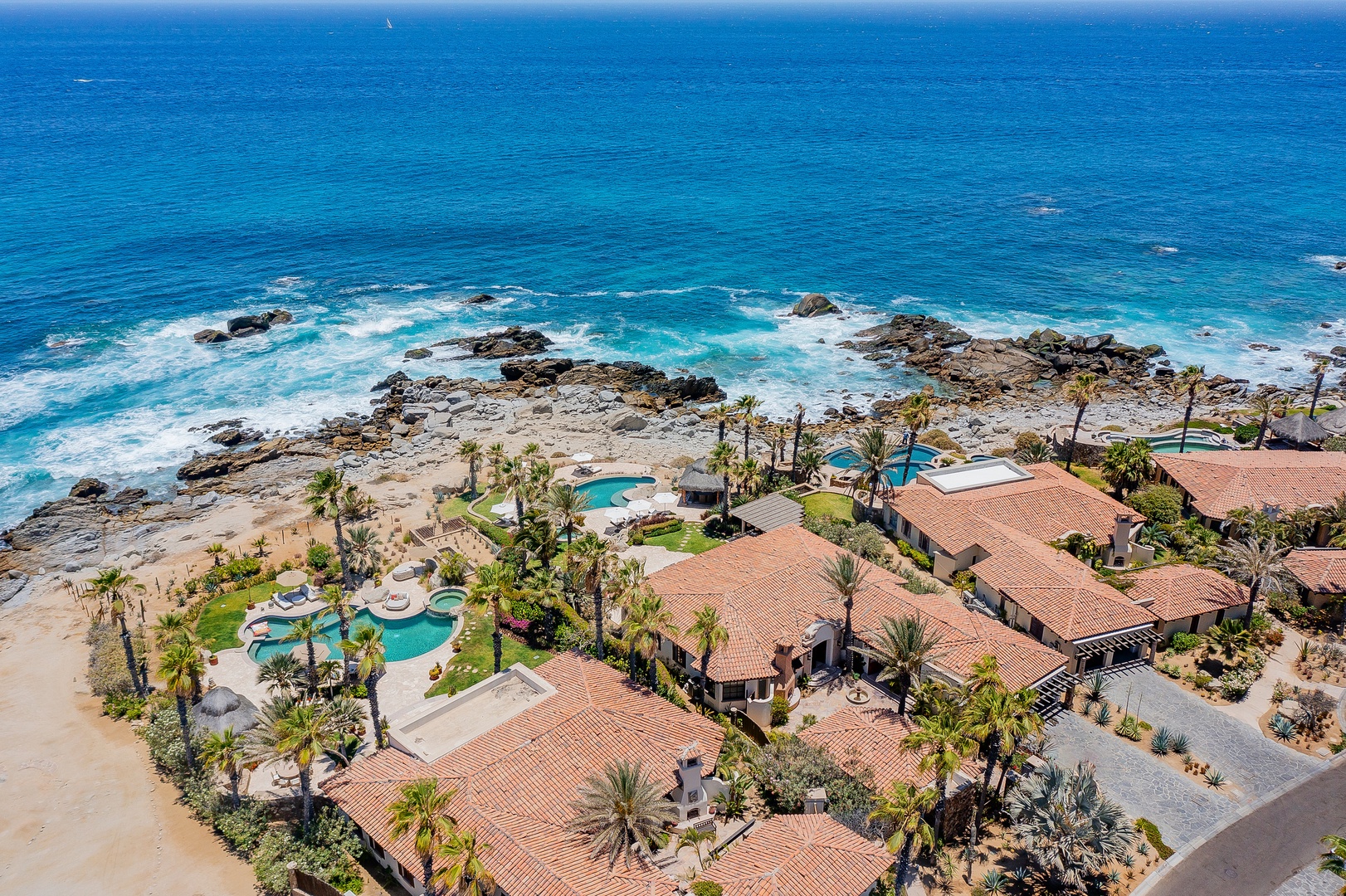 Beachfront resort complex featuring multiple villas with red tile roofs, swimming pools, and direct access to pristine coastline with dramatic rock formations.