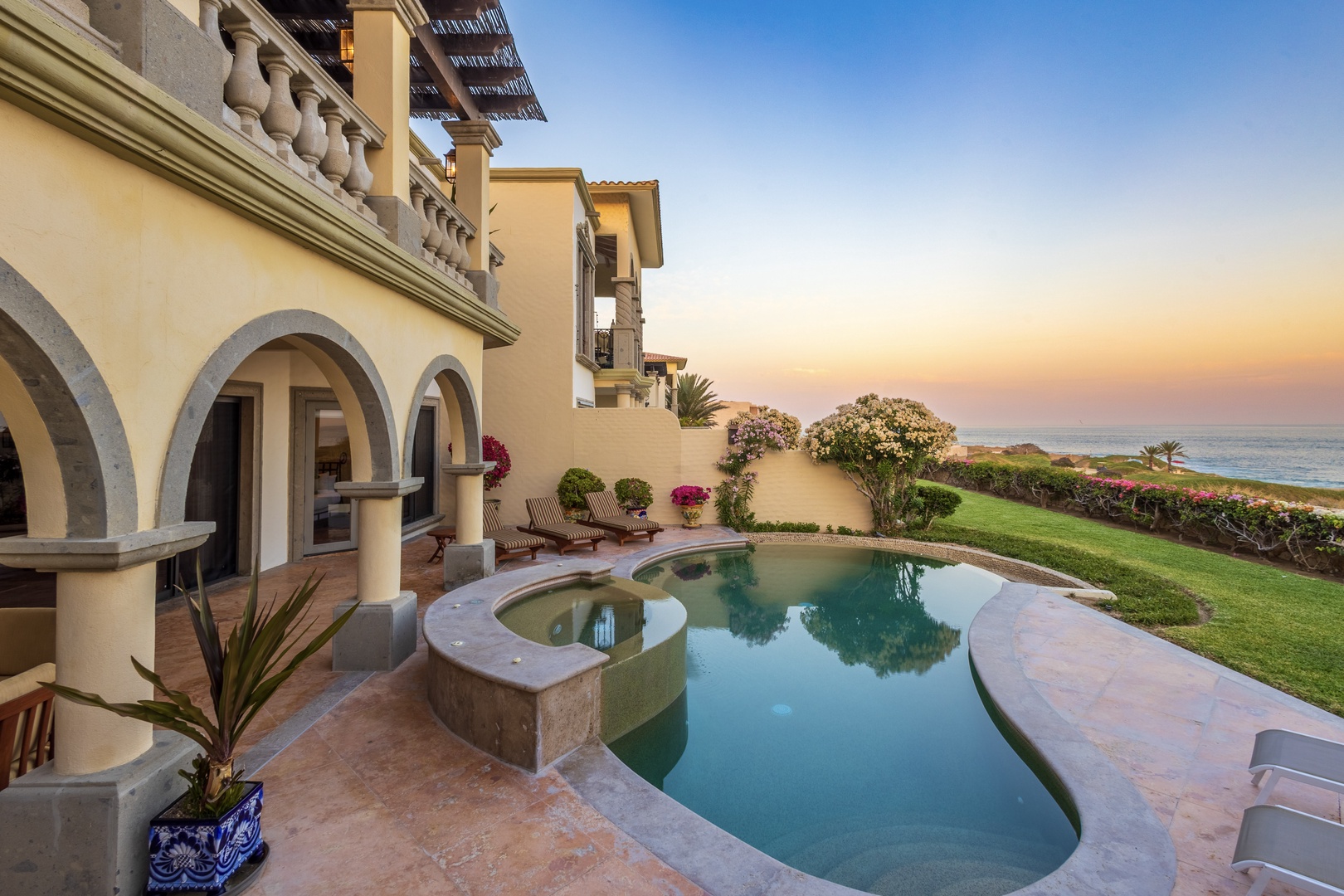 A luxurious beachfront villa with a stunning sunset backdrop. The property features Mediterranean-style architecture with arched columns, a private pool with integrated spa, and lush oceanfront gardens.