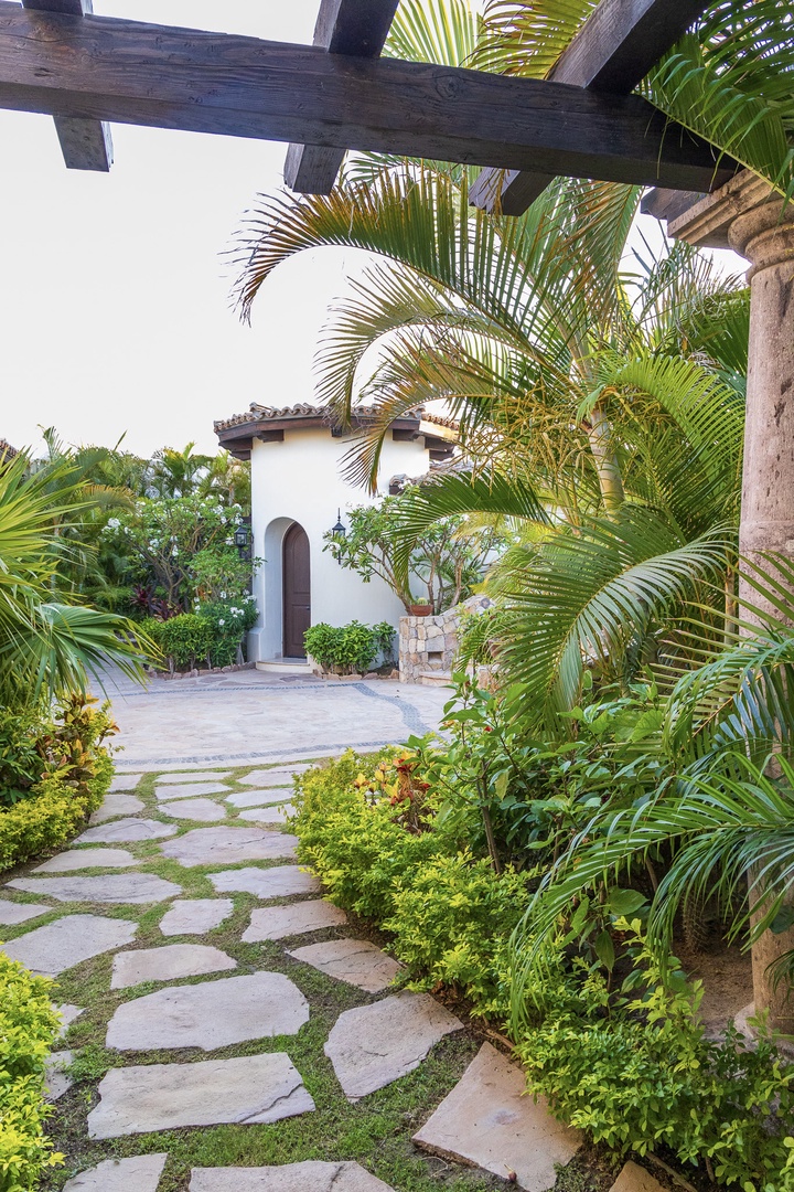 A stone walkway weaves through lush tropical gardens framed by a rustic pergola, guiding you to a charming arched entrance that invites quiet relaxation and coastal elegance.