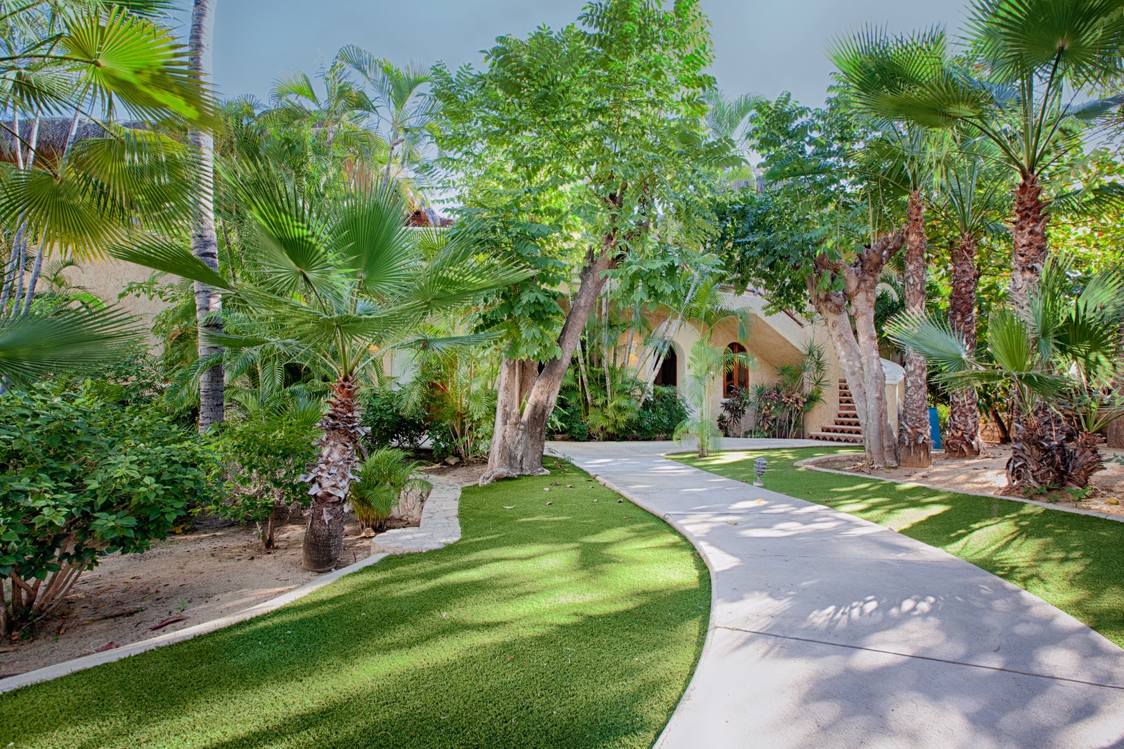 A winding pathway leads through lush tropical gardens with towering palms and manicured lawns toward the property entrance.