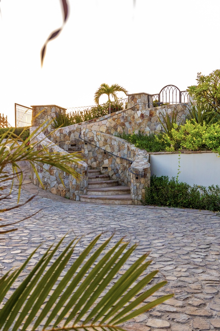 Stone stairway winds through lush tropical landscaping with cobblestone courtyard, leading to upper terrace with decorative ironwork, palm trees, and golden-hour ambiance.