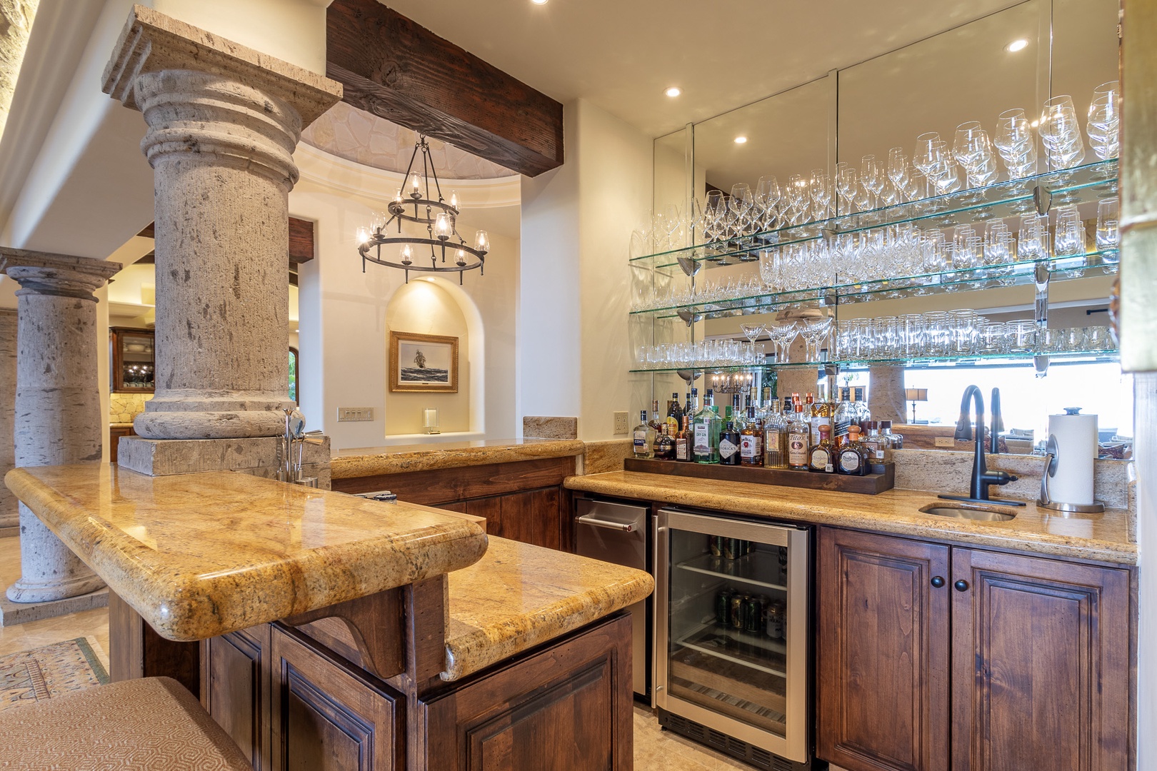 Your private bar features elegant glass shelving, premium spirits, and a granite counter with bar seating—perfect for crafting cocktails under rustic wooden beams and limestone columns.