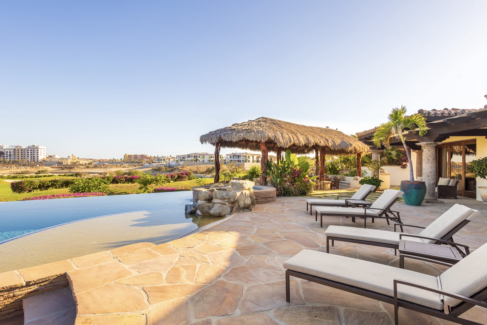 This expansive poolside terrace overlooks vibrant green grounds and distant resort skyline, featuring sunloungers, thatched palapas, and an infinity pool for peaceful lounging.