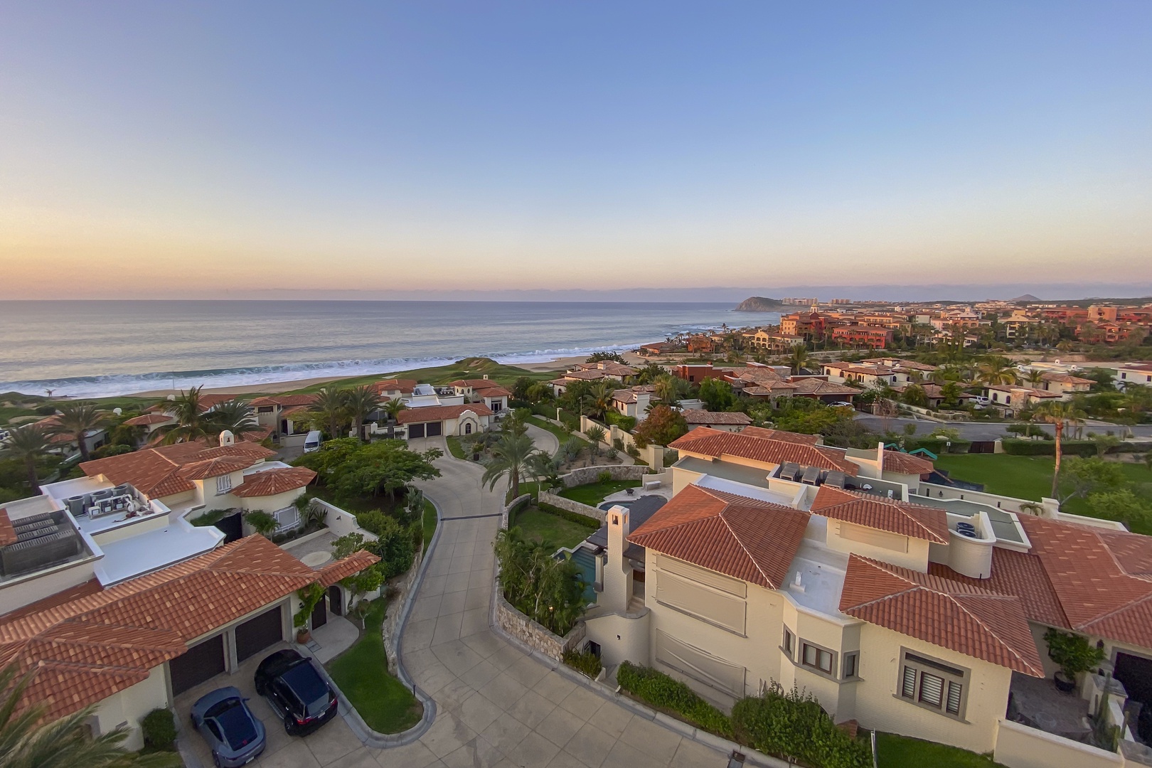 Elevated aerial perspective of a coastal residential community at twilight, showcasing villa-style homes with terracotta roofs nestled between ocean and town, capturing the serene beachfront location and surrounding neighborhood.