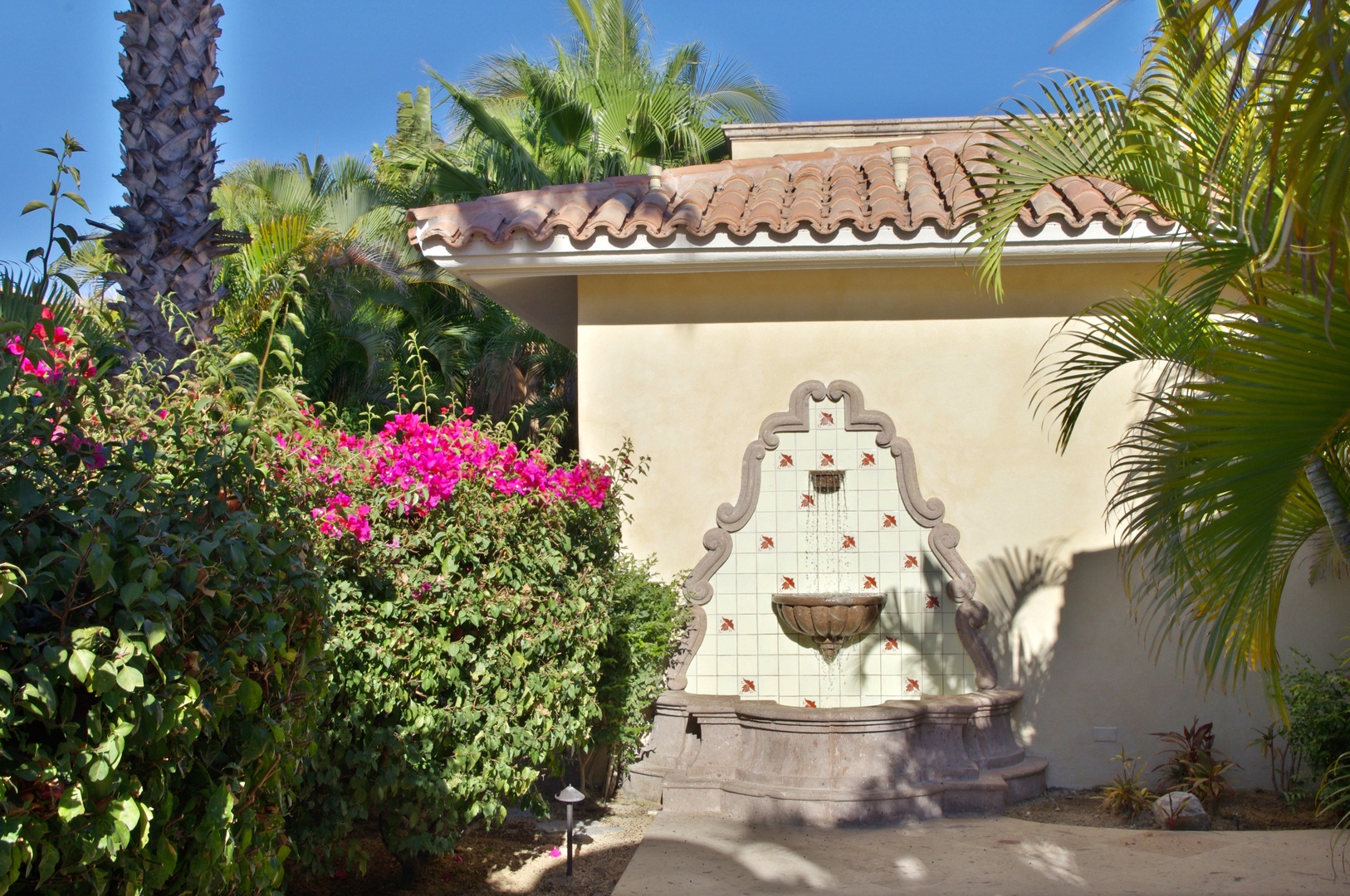 A decorative tiled fountain anchors the exterior courtyard, surrounded by lush palms and vibrant bougainvillea under terracotta roof tiles. The property's Spanish-style architecture creates a welcoming tropical ambiance.