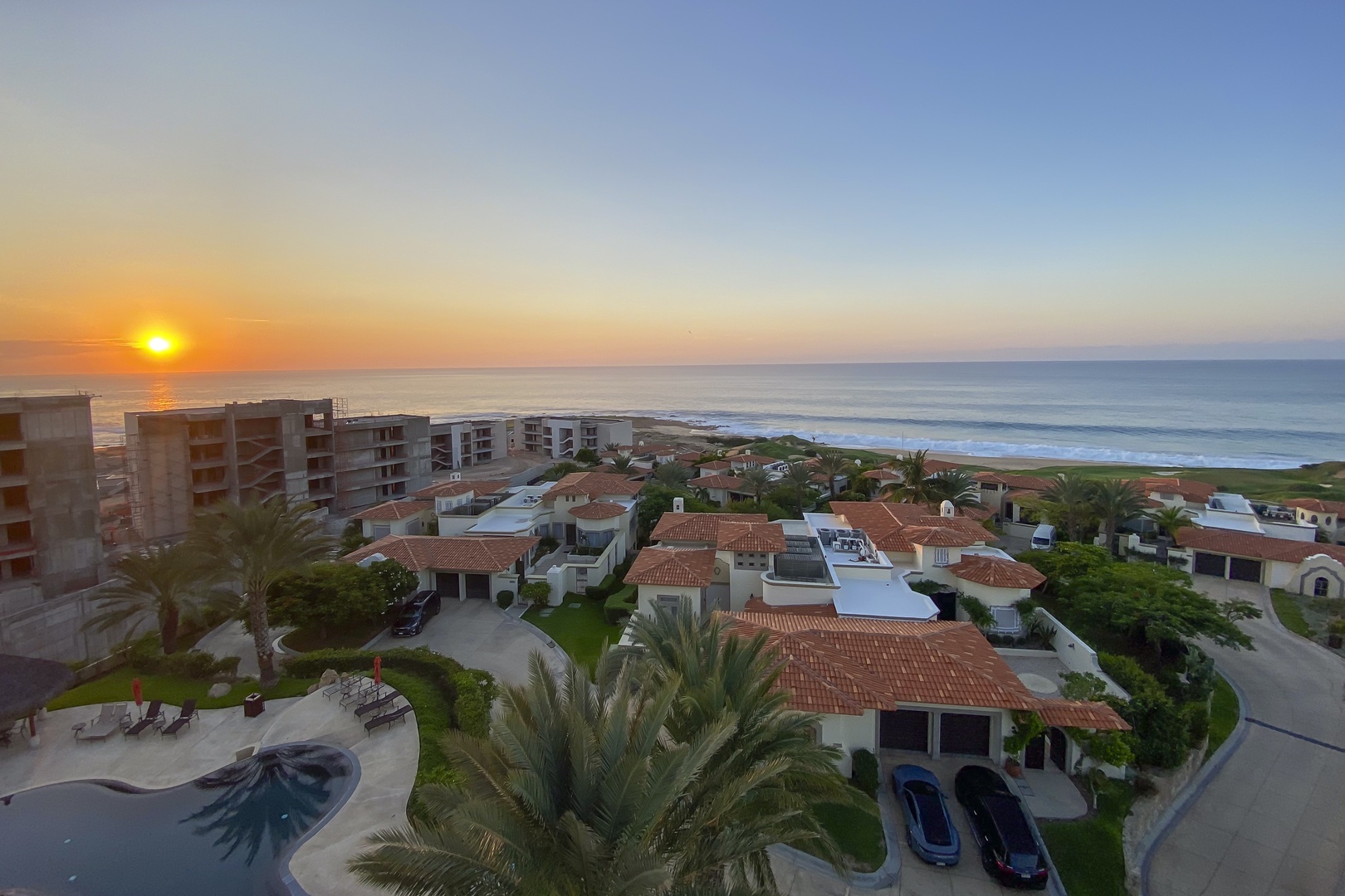 Aerial view of beachfront property showcasing coastal residences with terracotta roofs, palm trees, and the ocean stretching toward a golden sunrise horizon.
