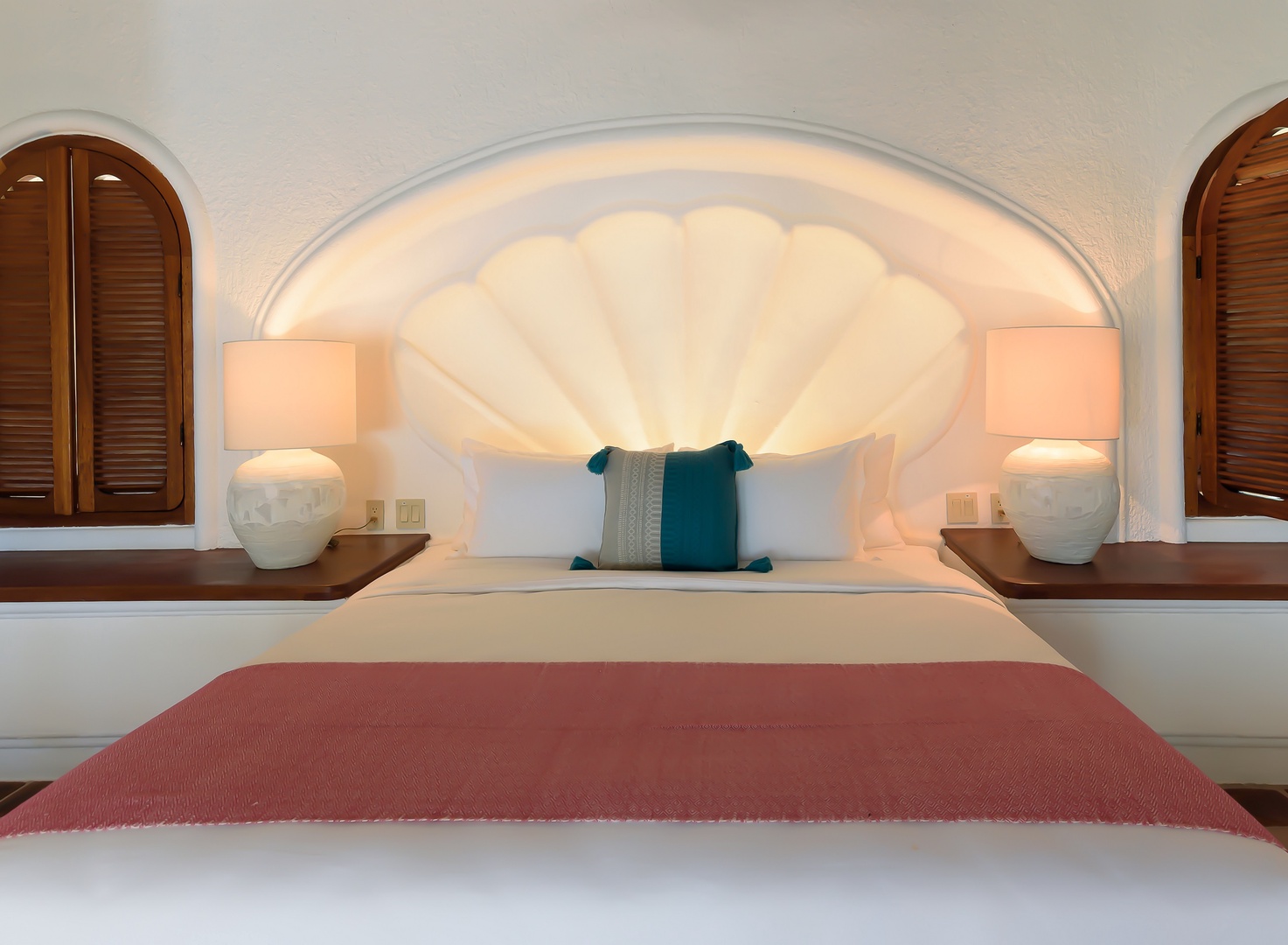 Sink into your plush bed framed by a stunning shell-shaped headboard, flanked by warm lamps and elegant shuttered windows that create your serene coastal-inspired retreat.