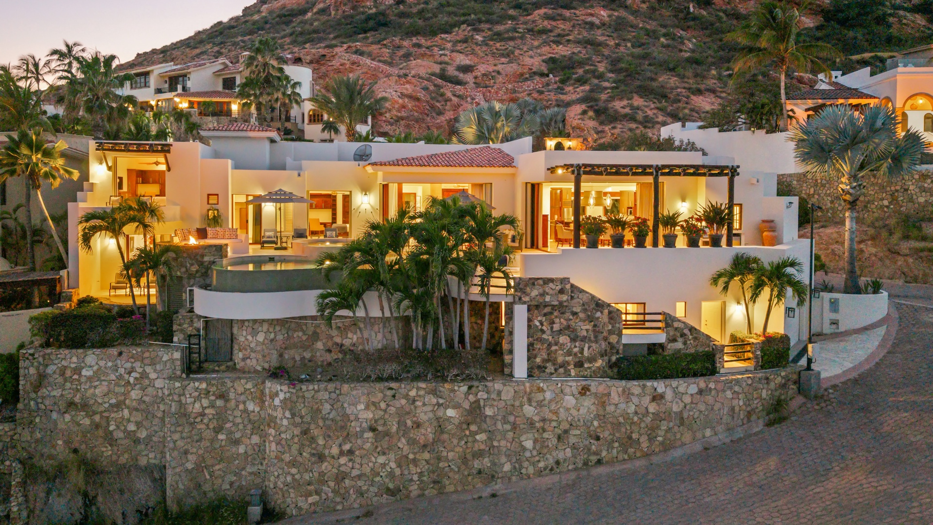 Luxurious hillside villa with stunning architecture and tropical landscaping illuminated against desert mountains at twilight.