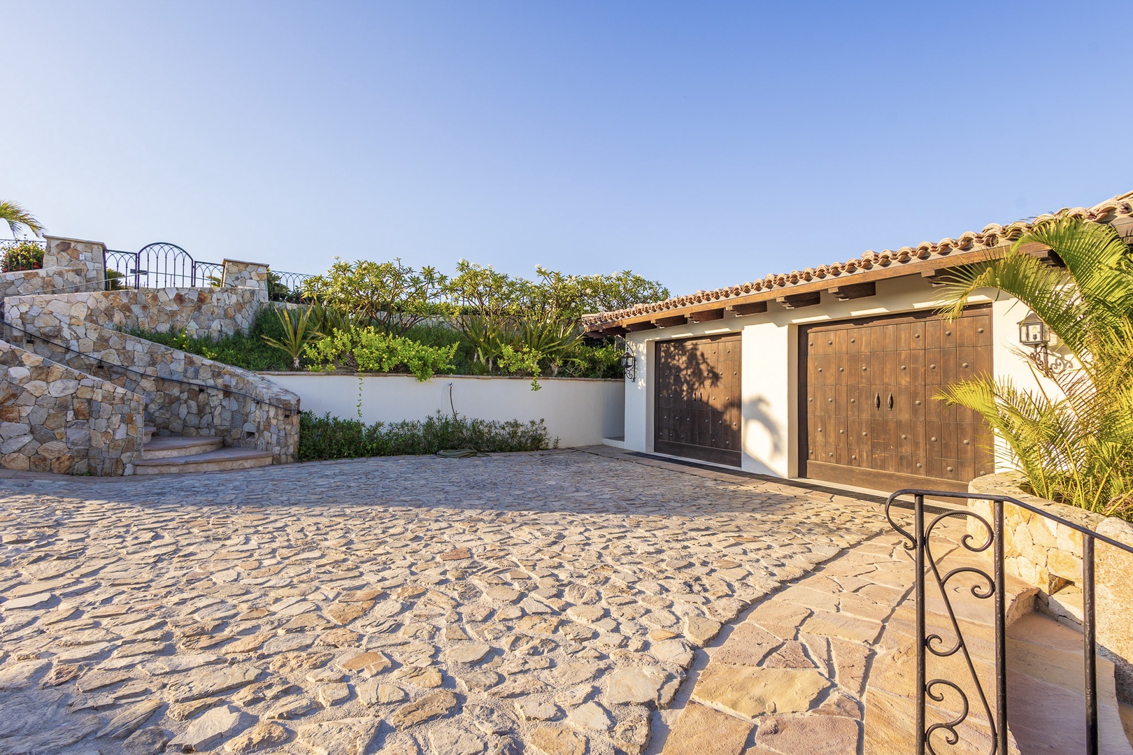 Charming stone courtyard entrance with Mediterranean-inspired architecture, clay-tiled roof, and wooden garage doors framed by lush tropical palms and greenery.