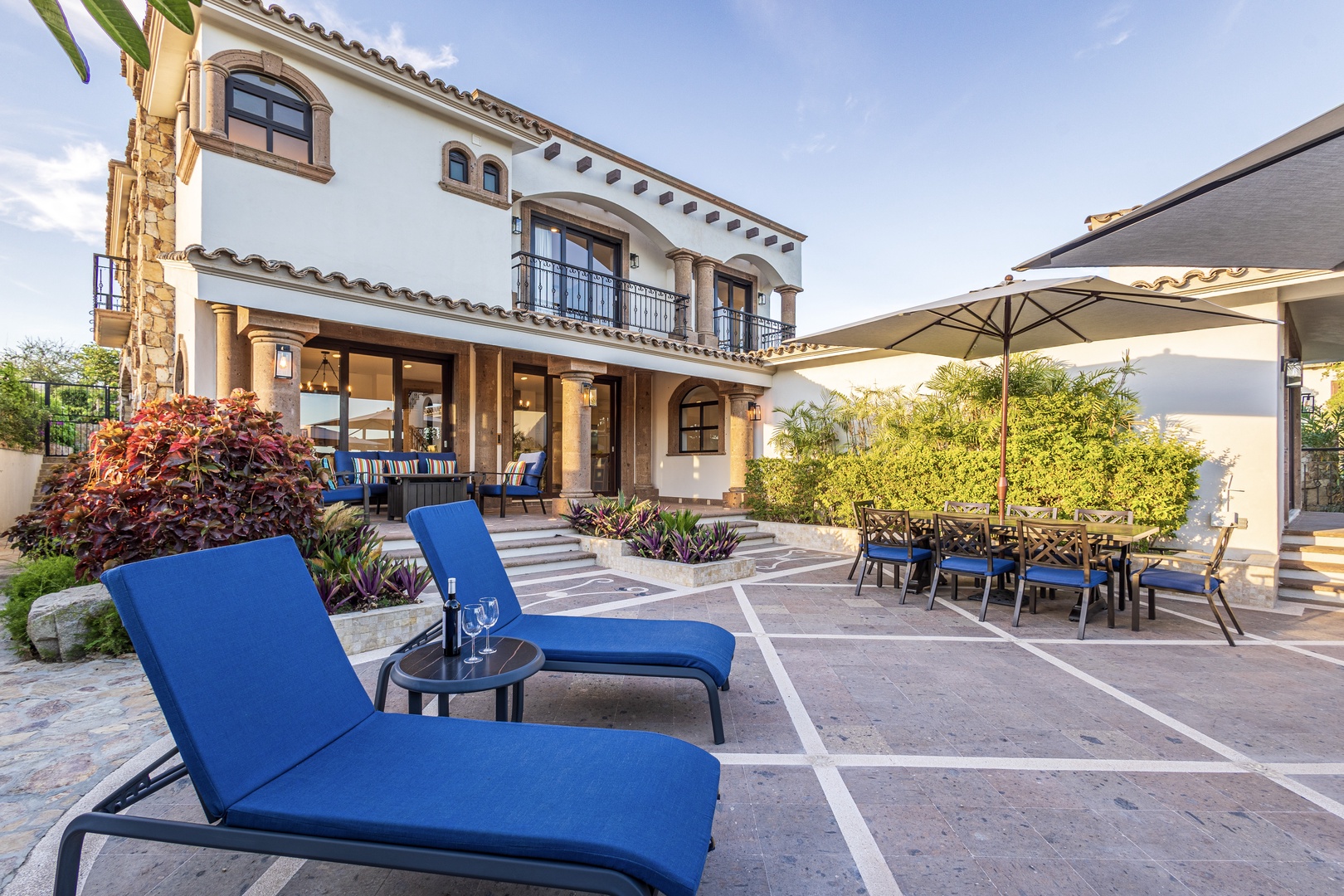 This stunning Mediterranean-style villa features a spacious patio with vibrant blue loungers and shaded dining—your perfect spot to unwind under the sun or enjoy alfresco meals surrounded by lush landscaping.