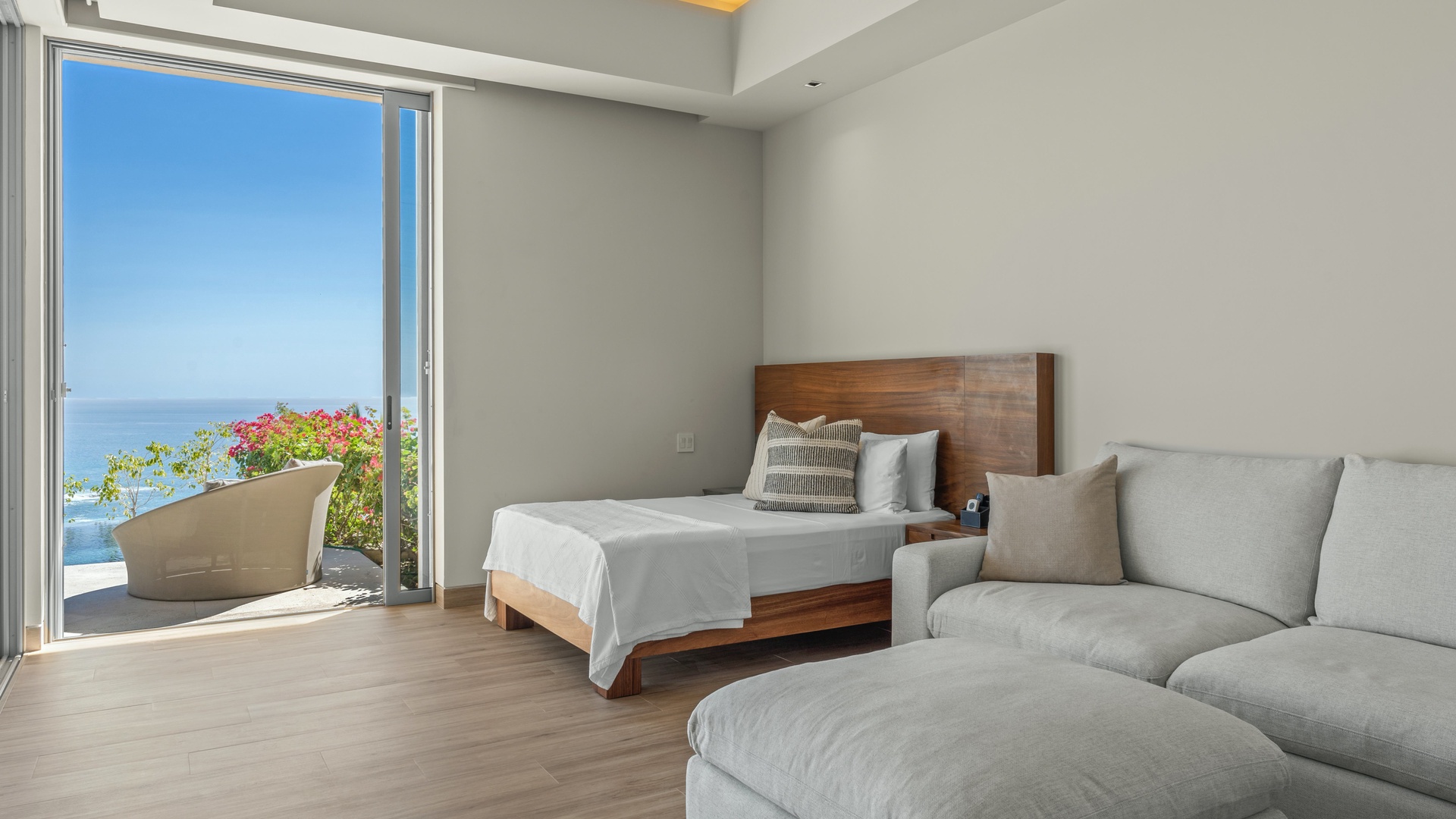 Step into your serene retreat where a comfortable bed and plush lounge seating invite relaxation, while open doors reveal a stunning ocean view and vibrant blooms right outside.