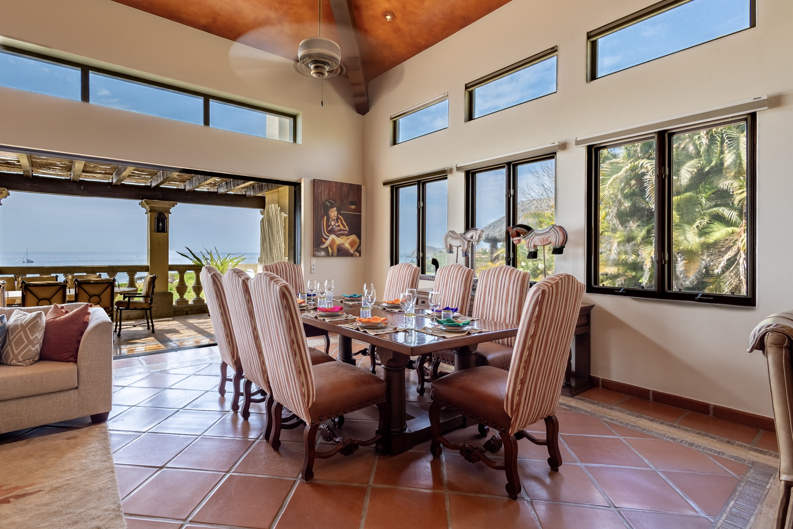 Your private dining retreat features soaring ceilings with warm wood tones, large windows framing ocean and palm tree views, and elegant seating for memorable meals together.