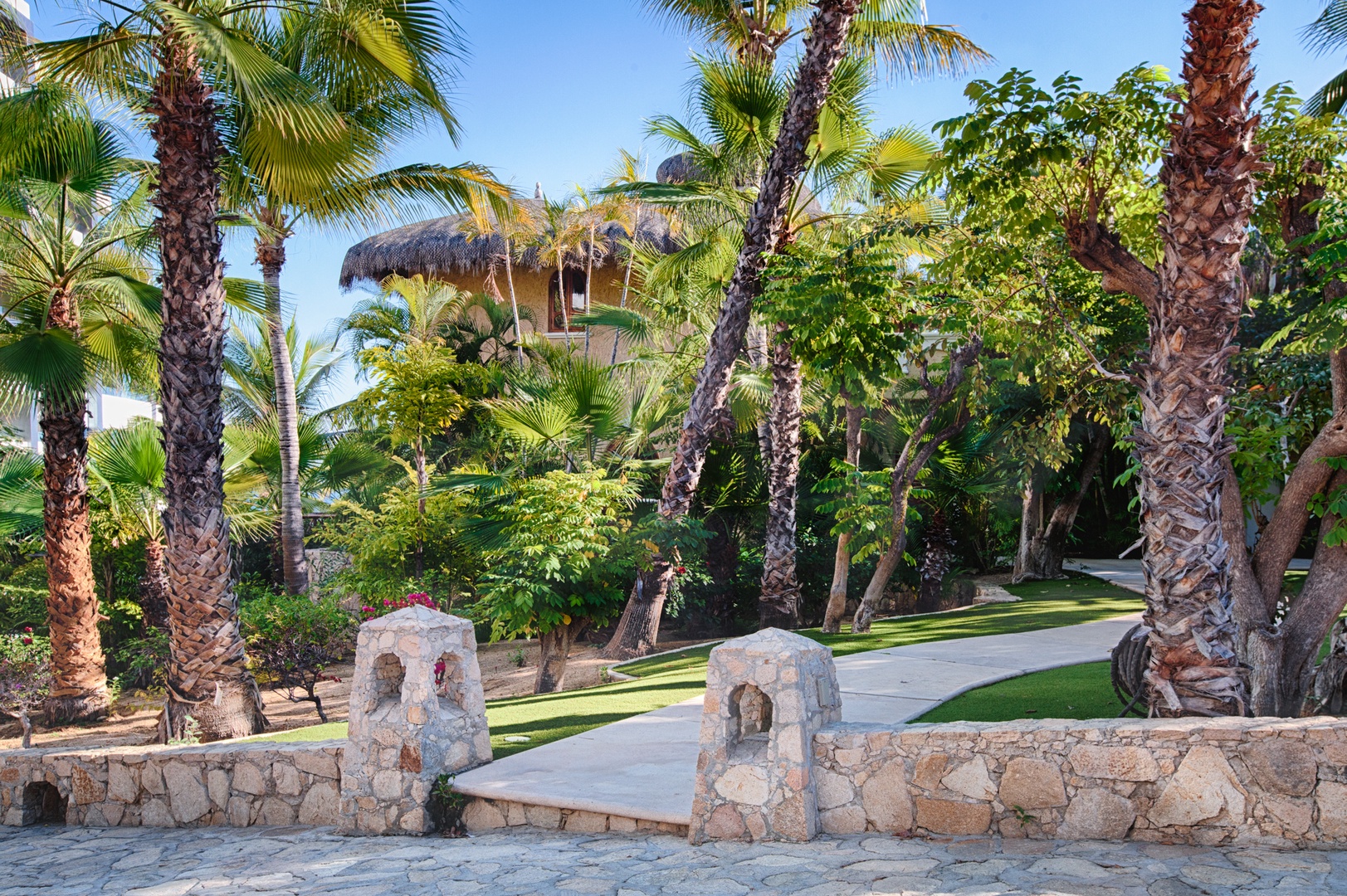 A tropical resort setting with stone pillars framing pathways through lush palm trees and landscaped grounds.
