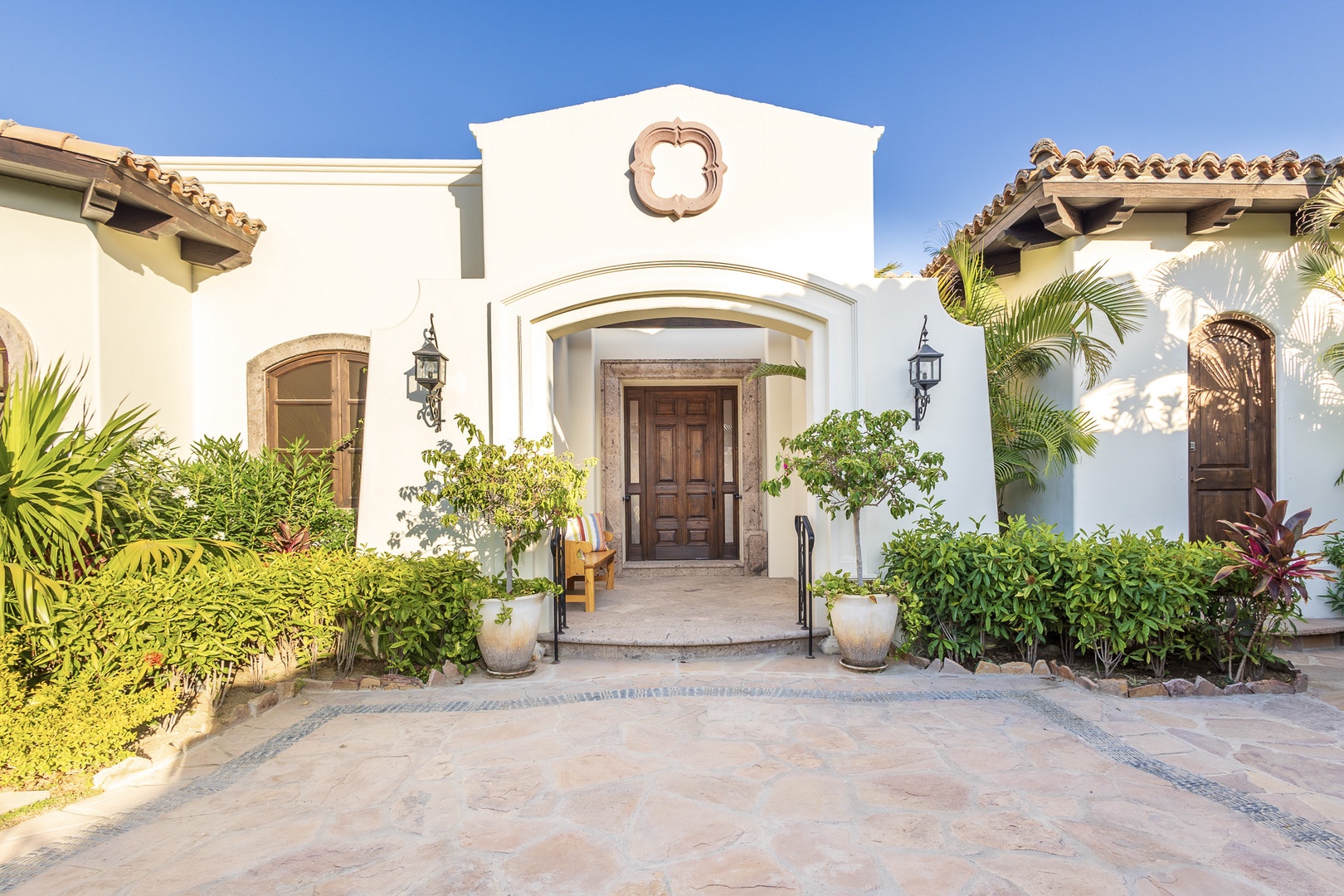 A stunning Spanish-style villa with arched entryway and terracotta roof welcomes guests through lush tropical gardens and elegant cobblestone courtyard.