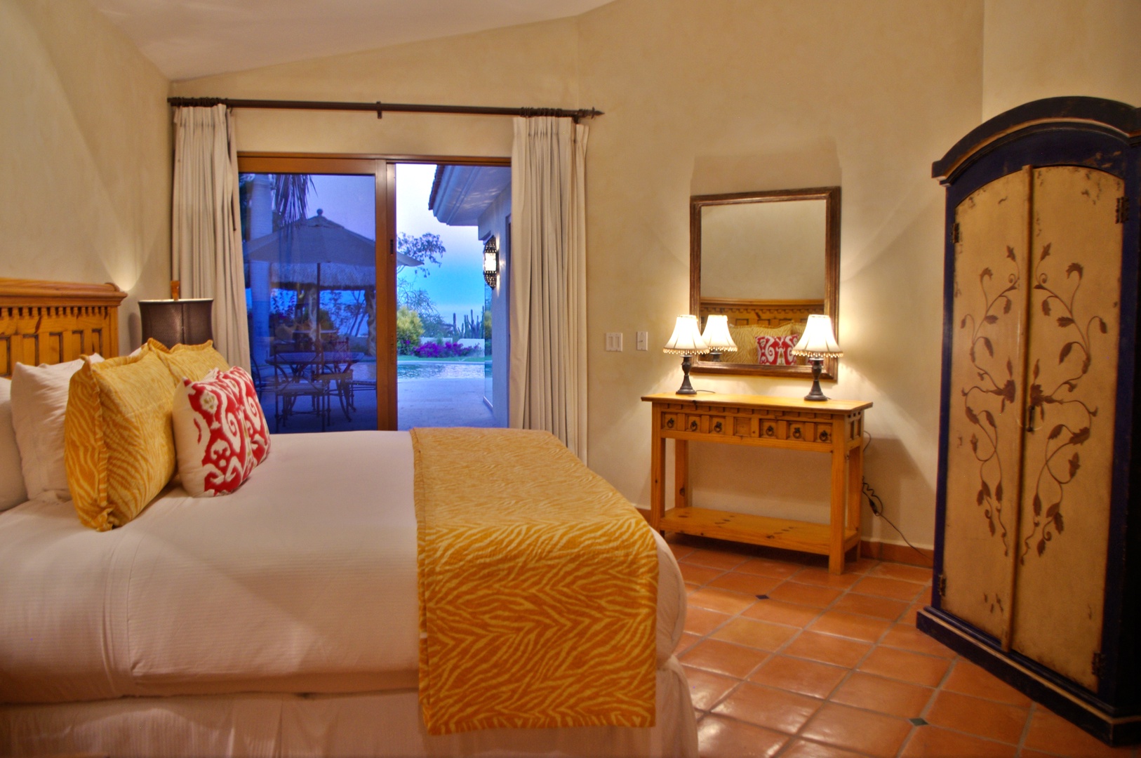 Your bright bedroom offers comfort and charm with a golden bedspread, tropical patio views, and handcrafted wood furniture. Step outside to your private terrace with pool glimpses.