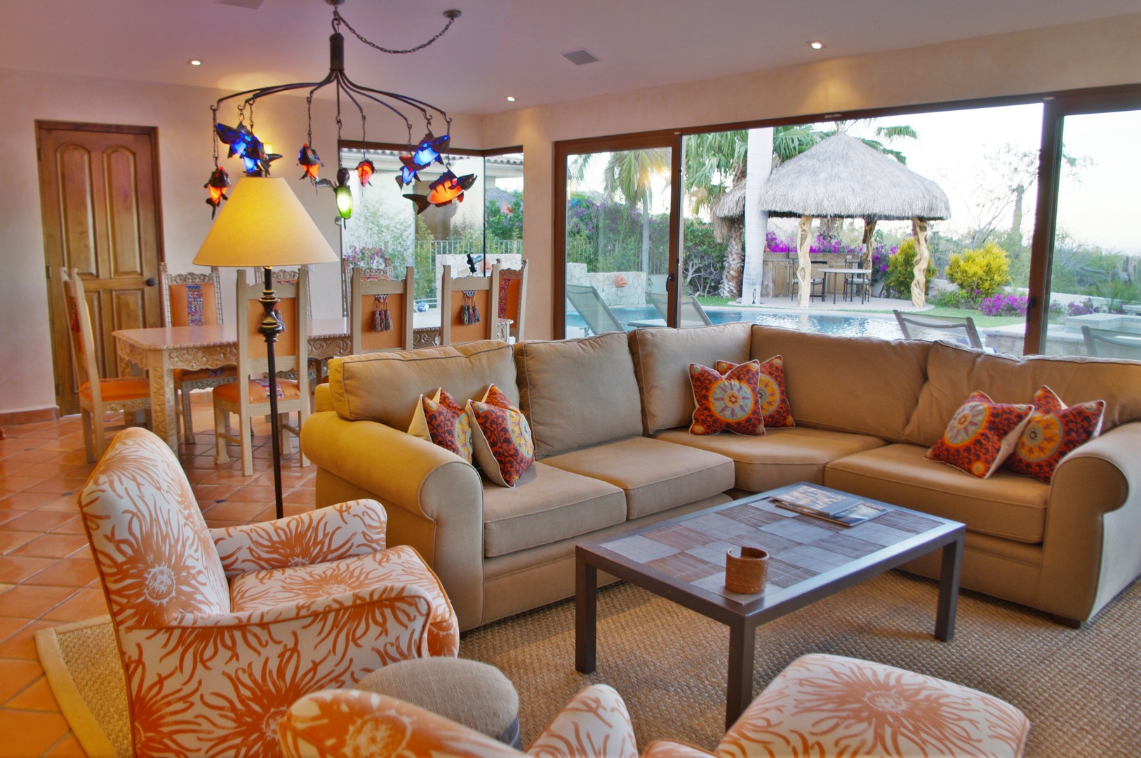 Settle into your sun-filled living space where plush seating and warm tropical tones invite you to unwind. Through expansive windows, your private pool and palapa await beneath swaying palms.