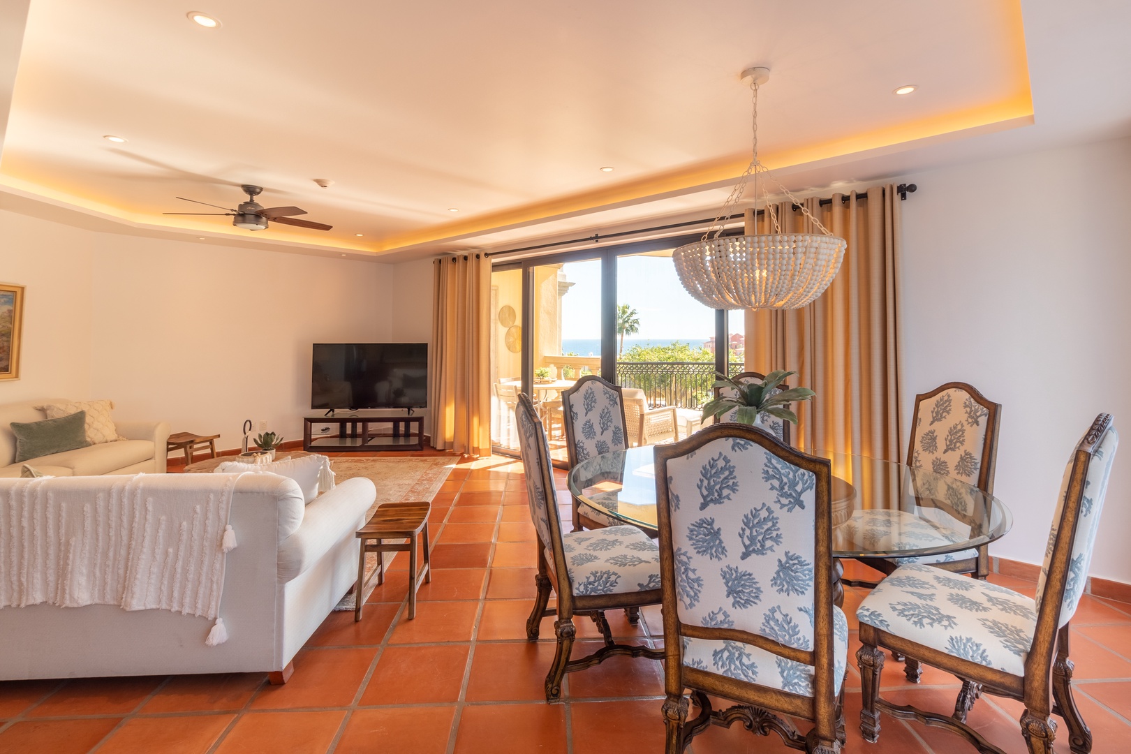 Step into your sunlit haven where warm terra-cotta tiles and elegant furnishings create an inviting atmosphere. The open layout flows from the plush living area to the dining space, while ocean glimpses through sliding glass doors add calm.