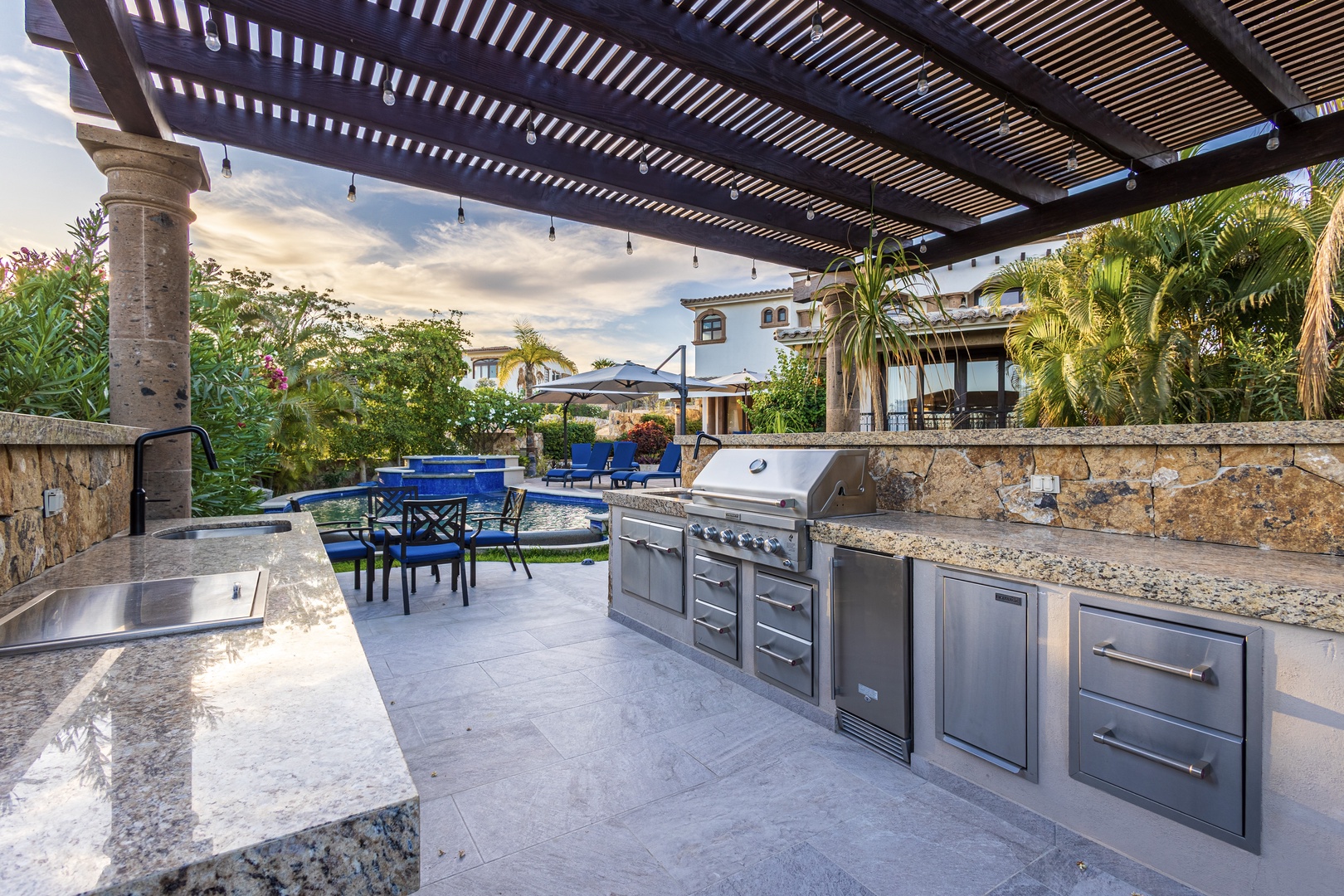 This outdoor kitchen features a full-size grill, stainless-steel appliances, and granite countertops—perfect for cooking while enjoying tropical breezes, lush greenery, and views of the sparkling pool and villa beyond.