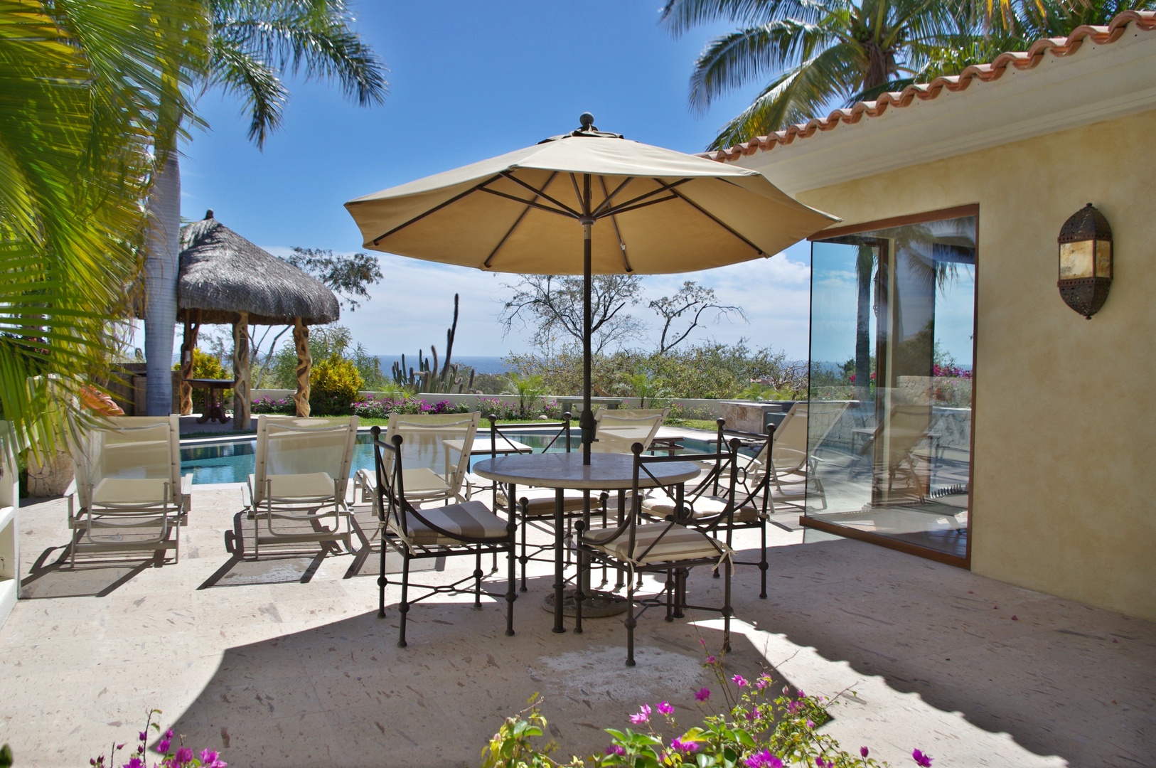 Relax poolside at your private outdoor dining area, shaded by palms and overlooking the sparkling pool and distant sea views—a perfect spot for leisurely meals under clear blue skies.