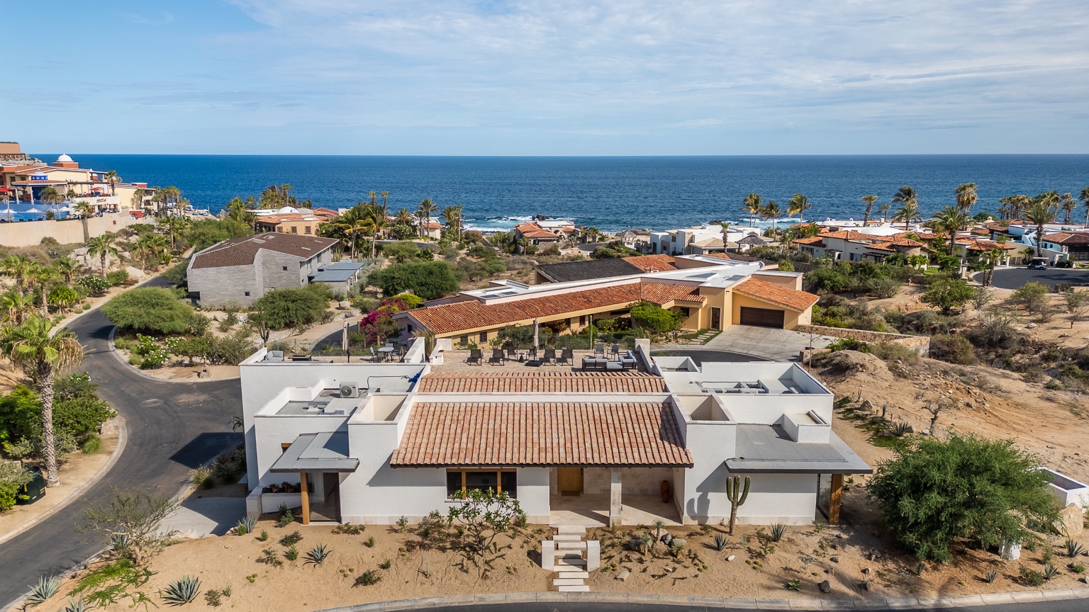 Elevated coastal property featuring contemporary architecture with tile roofing, positioned on a hillside overlooking the ocean and neighboring residential area.