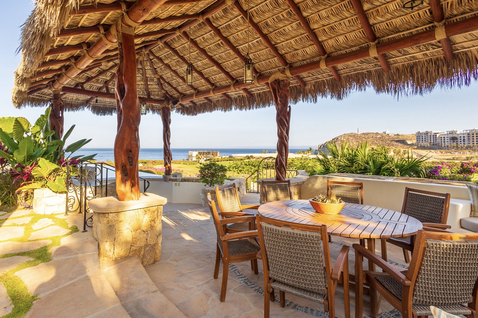 A stunning coastal terrace featuring a traditional palapa with woven palm roof, offering sweeping ocean views and distant hillside vistas. The outdoor dining area is perfect for seaside meals.