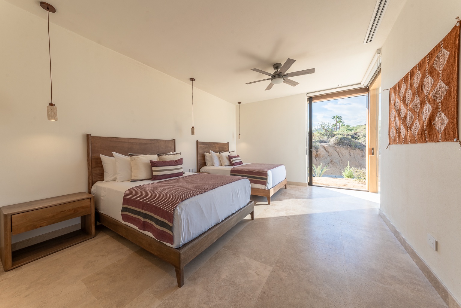 Wake up in your bright desert suite with two cozy beds, a modern ceiling fan, and stunning natural light framing views of cacti and native plants just outside your door.