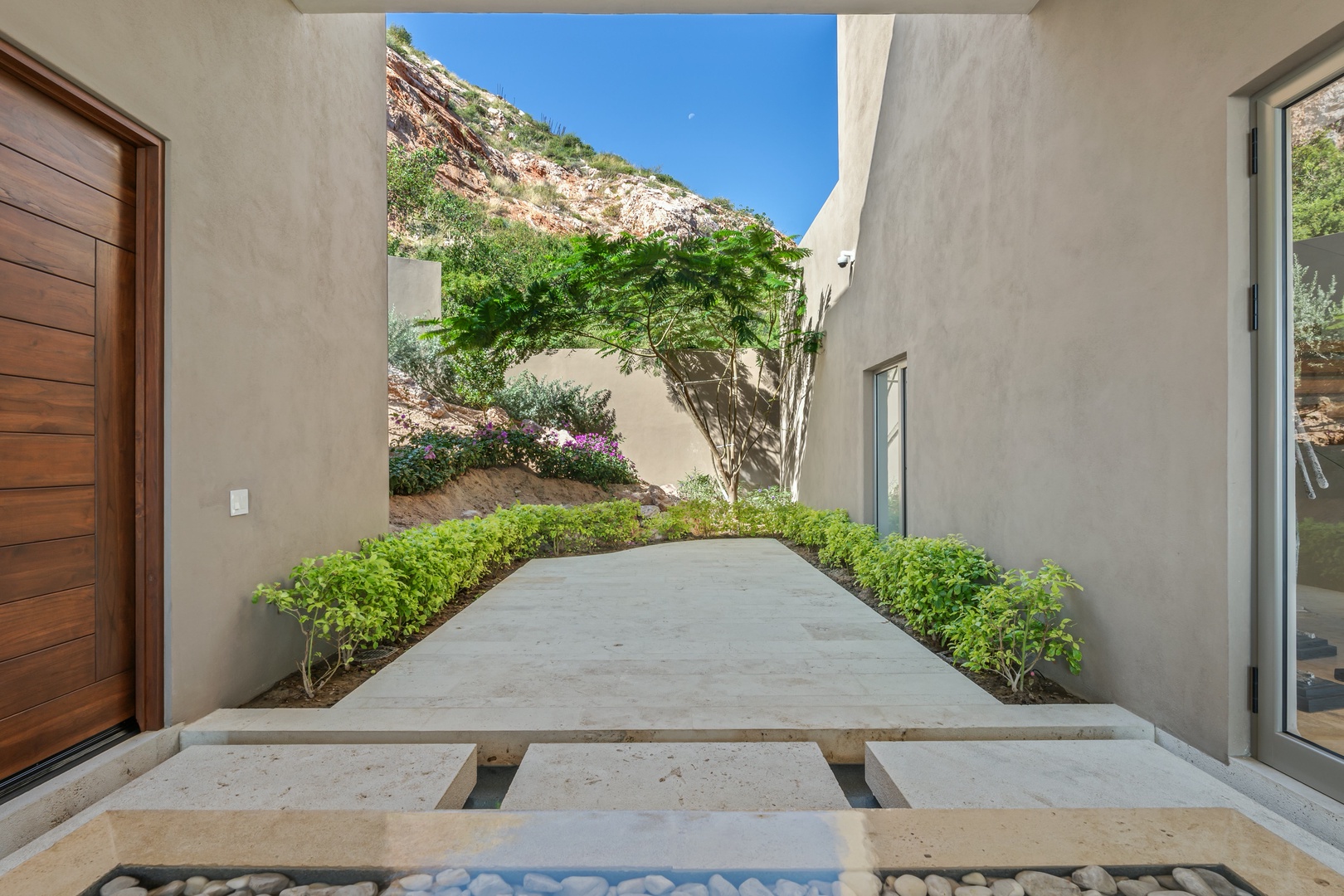 A modern courtyard entrance framed by native desert plantings and a rocky hillside, offering a serene arrival experience to the property.