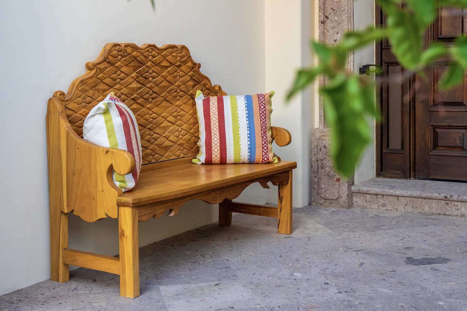 Charming wooden bench with traditional carved backrest and colorful cushions welcomes you to this property's entrance—a cozy spot to relax before stepping inside.
