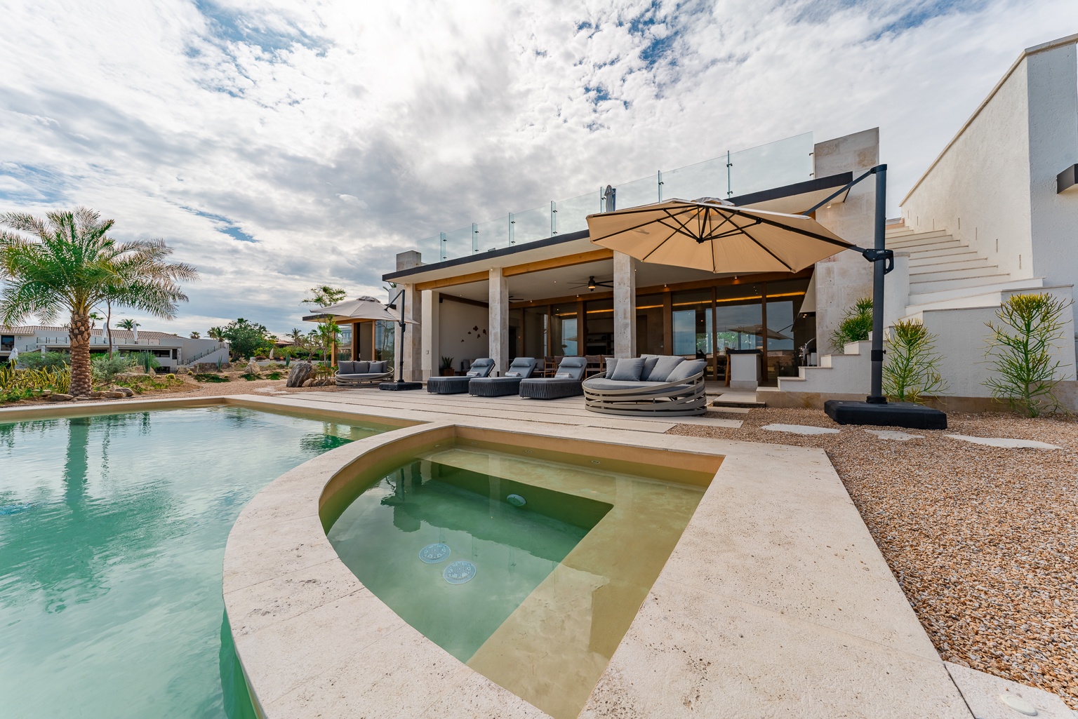 Modern villa exterior showcasing a serene pool area with integrated hot tub, shaded lounge seating under a large umbrella, and contemporary architecture framed by tropical palms and desert landscaping.