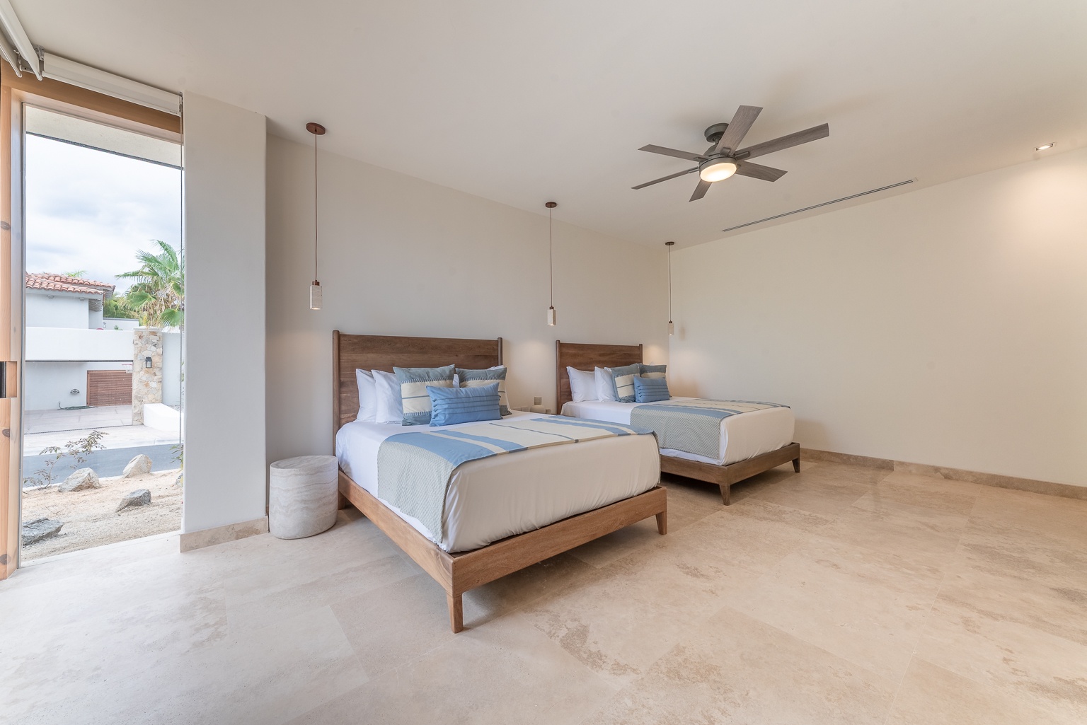 Your spacious bedroom features two beautifully made beds with coastal-inspired linens, modern pendant lighting, and a ceiling fan to keep you comfortable. Step outside to your private patio view.