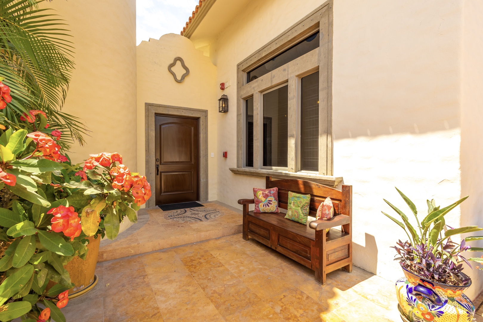 A welcoming entryway framed by sun-warmed walls and vibrant tropical blooms, with a handcrafted wooden bench inviting you to pause and soak in the serene courtyard ambiance.
