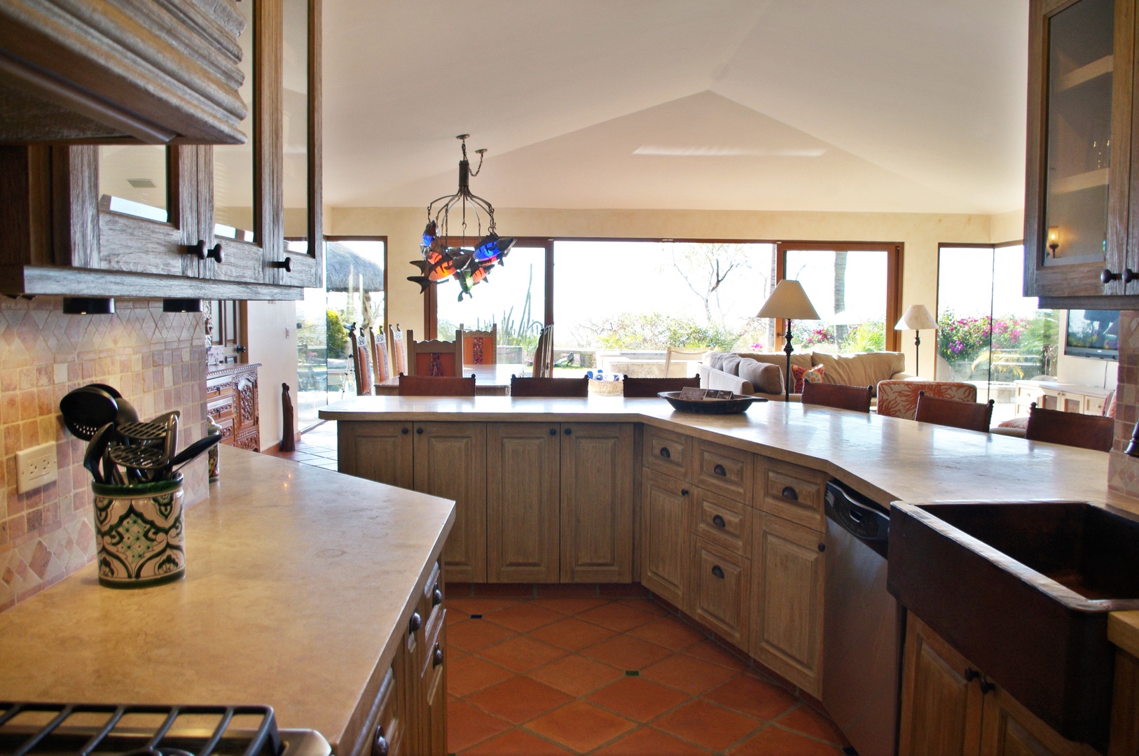 Your spacious kitchen flows seamlessly into a sun-drenched living room, where large windows frame stunning views and natural light dances across rich wood cabinetry and warm terracotta floors.