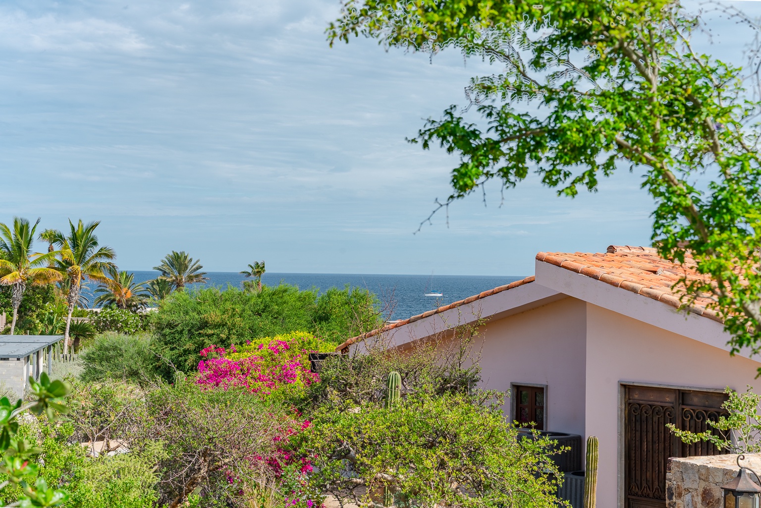 This charming villa features a terracotta tile roof and white stucco exterior, surrounded by lush tropical gardens with vibrant bougainvillea, offering stunning sea views beyond swaying palm trees.