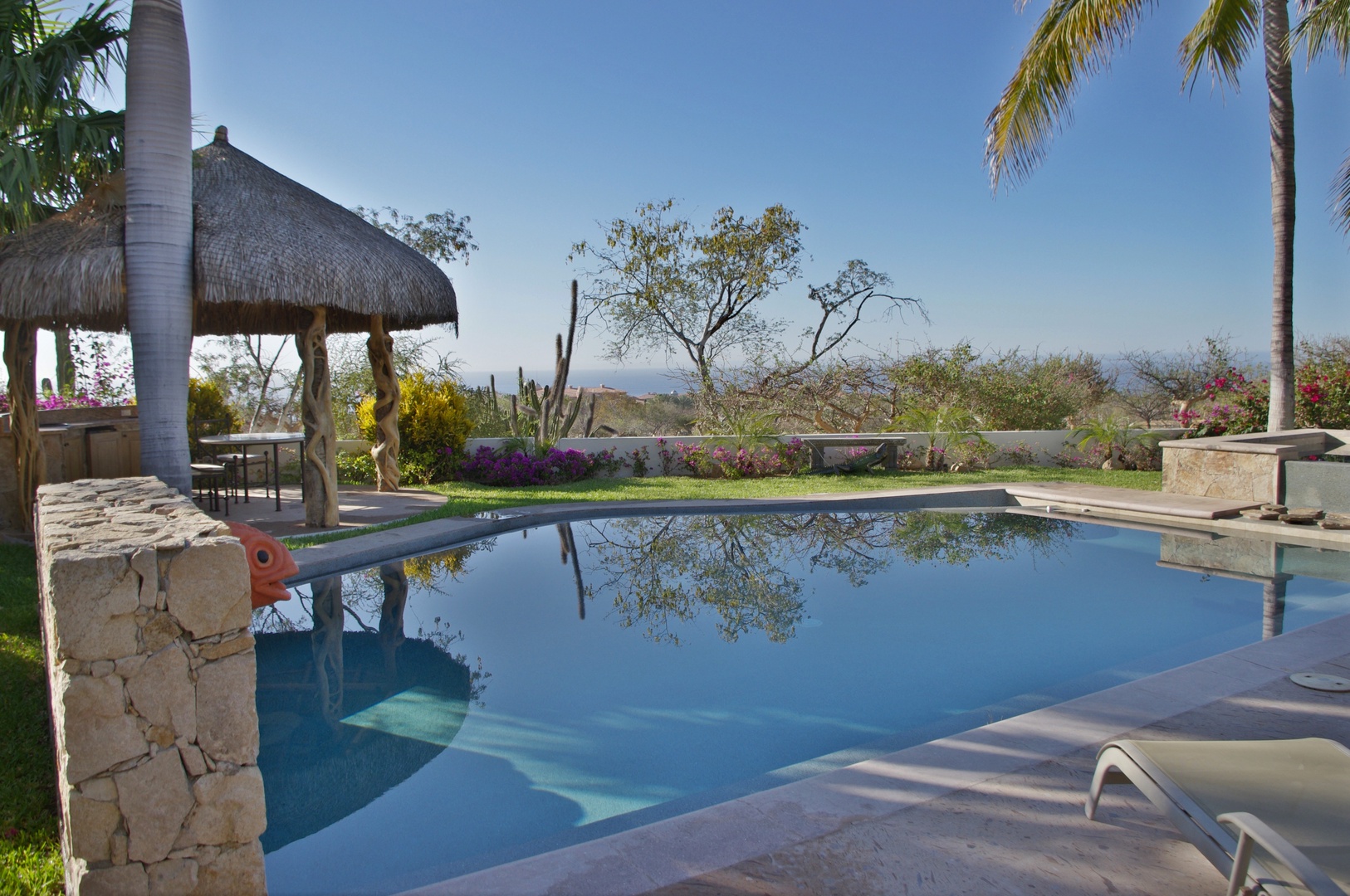 A tranquil pool area framed by palms and vibrant gardens, offering serene ocean views and shaded relaxation under a thatched palapa.