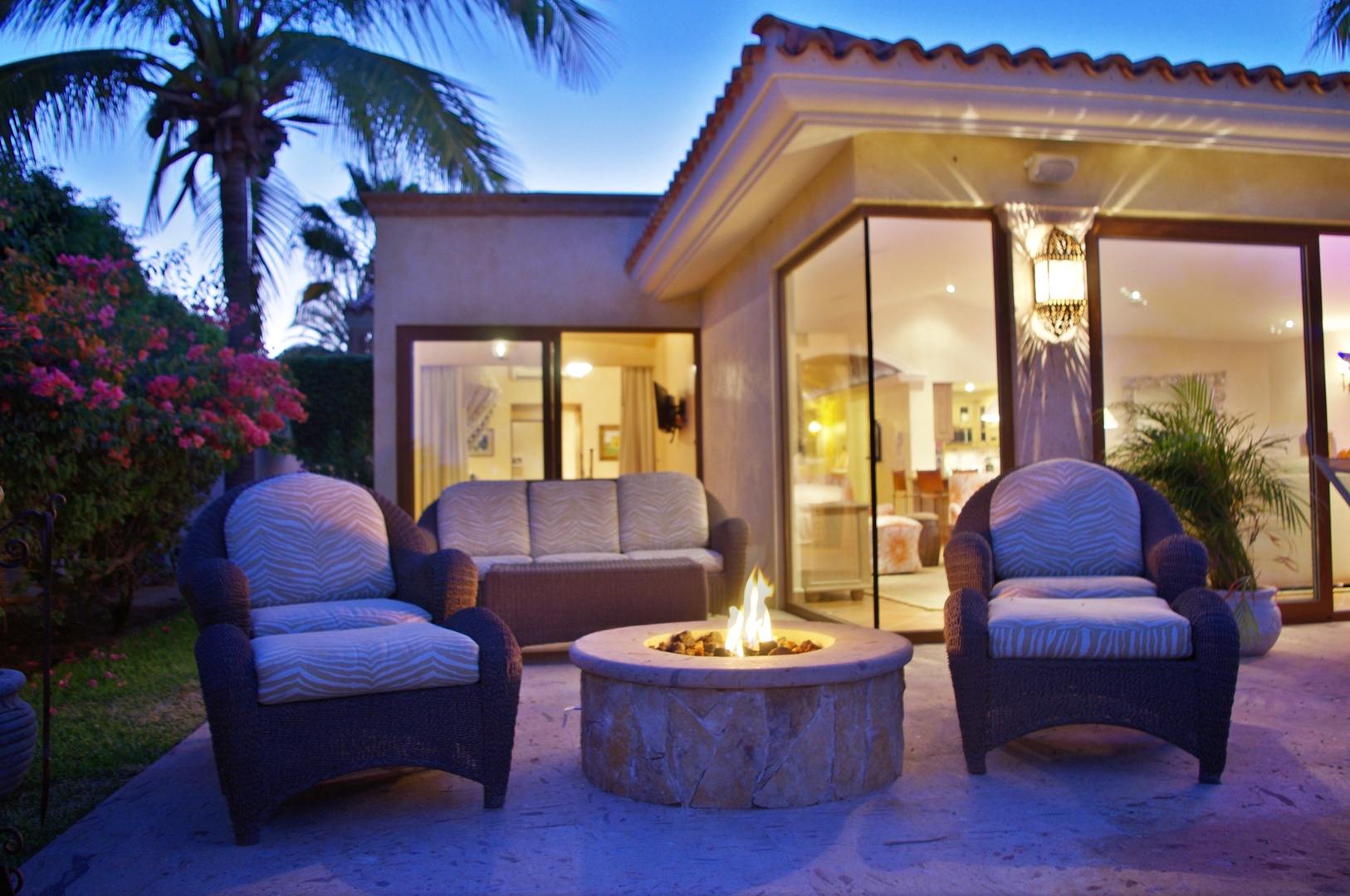 This inviting outdoor patio features a cozy fire pit surrounded by plush seating, perfect for relaxing under the tropical evening sky near your villa.