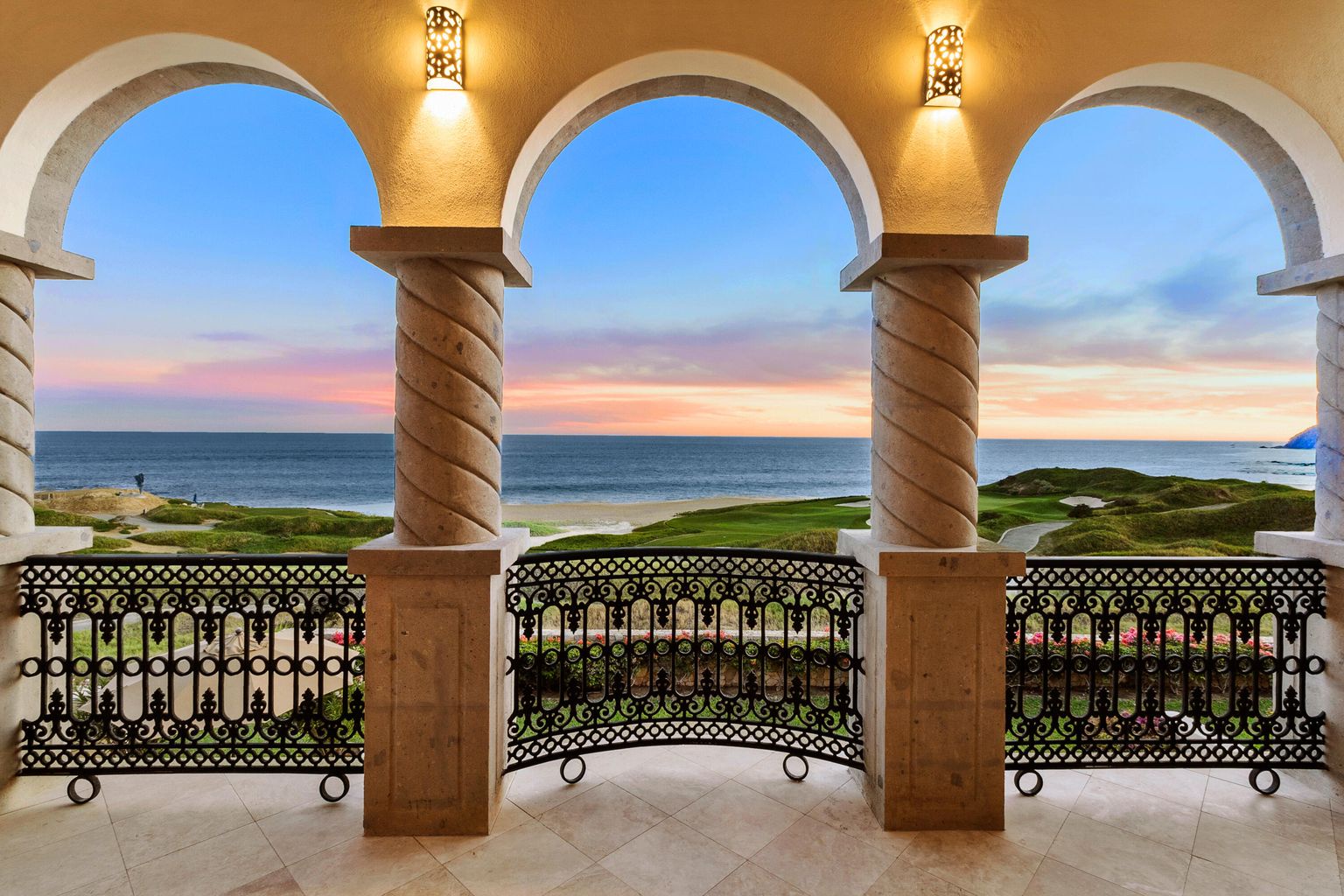 Stunning ocean views frame a pastel sky at dusk through elegant arches with intricate ironwork and warm lighting on your private terrace.