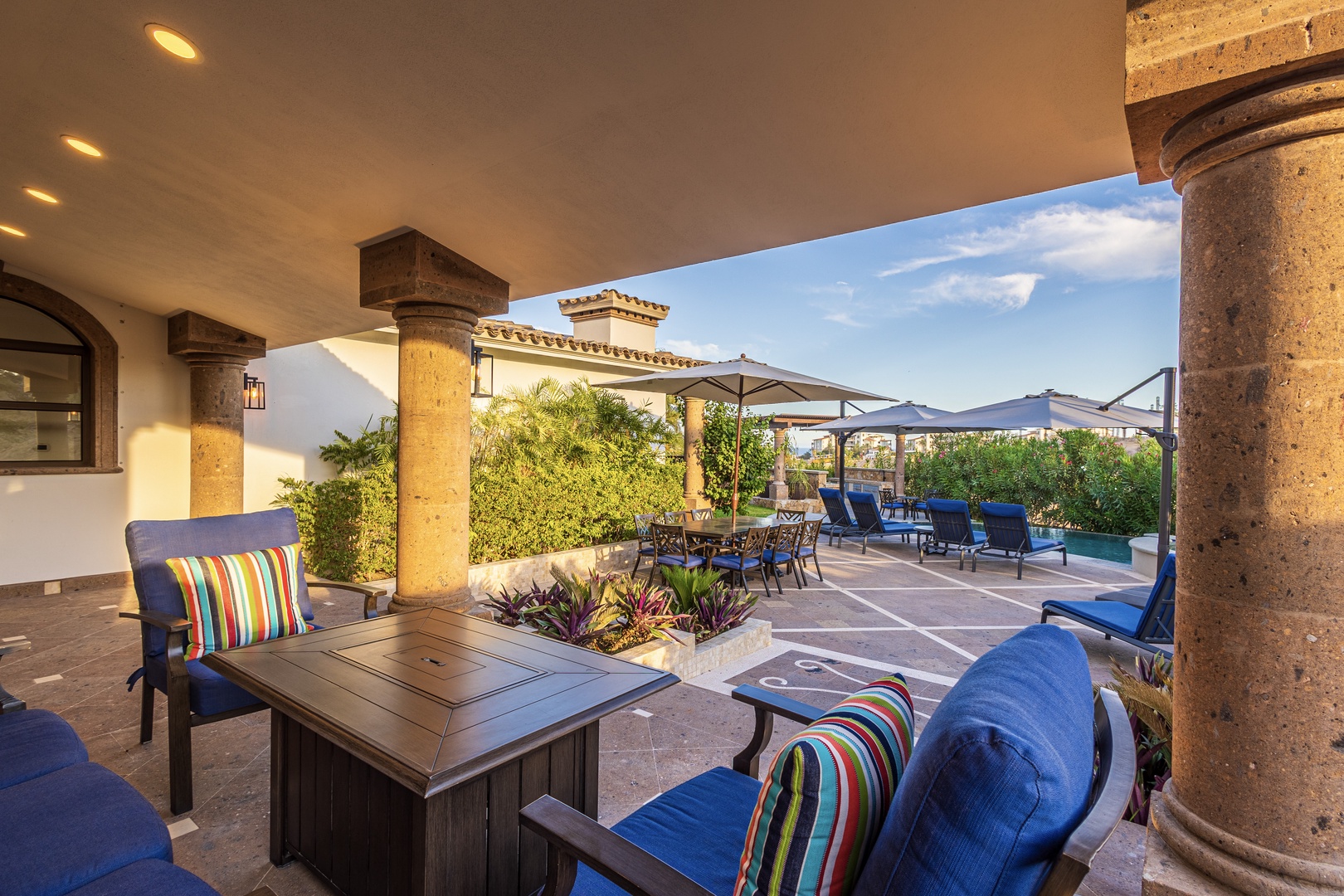 Your covered patio opens to a sunny courtyard with poolside seating, lush greenery, and vibrant blue chairs perfect for outdoor relaxation.