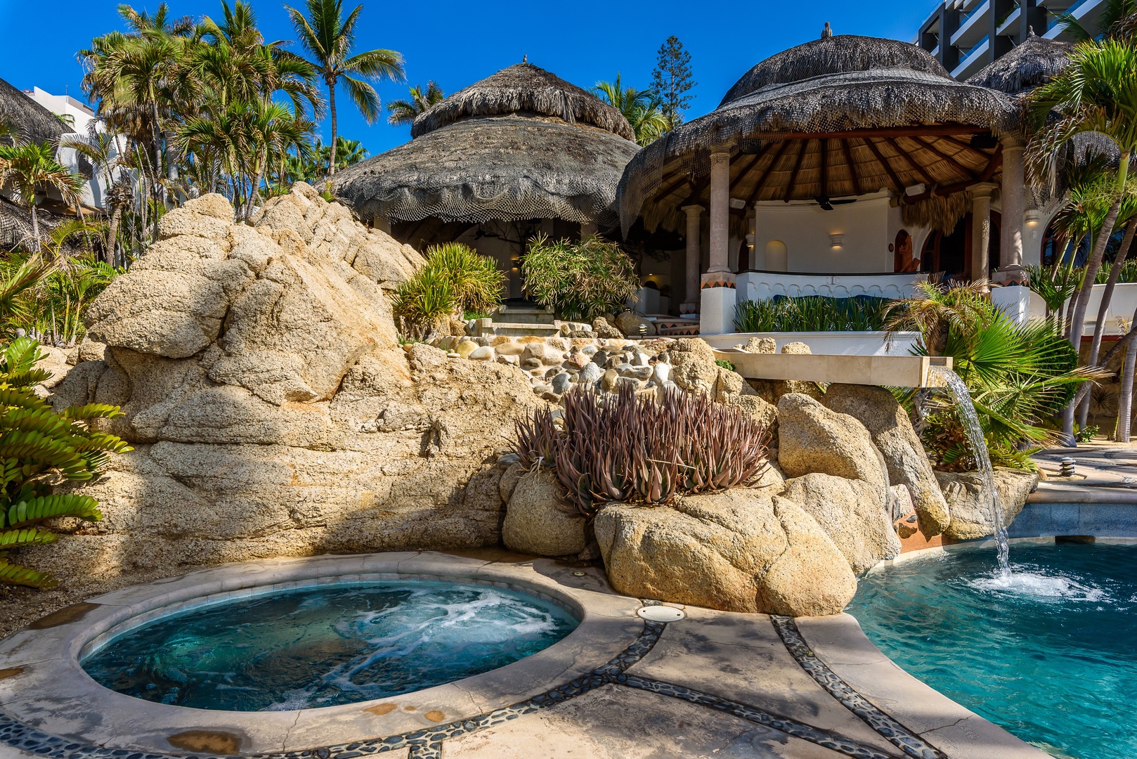 Soak in your private hot tub surrounded by natural rock formations and lush palms, with a thatched-roof cabana offering shade and relaxation steps away.