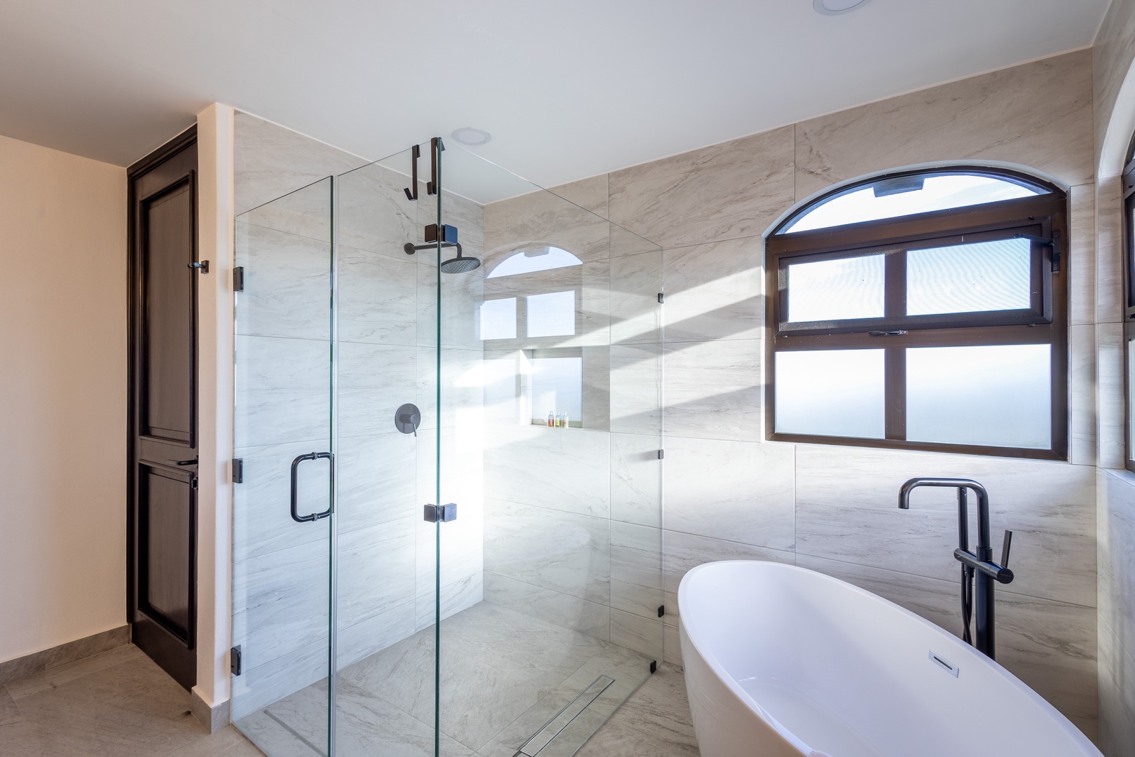 Unwind in your spa-like bathroom featuring a sleek soaking tub, spacious walk-in shower, and abundant natural light through the elegant arched window.