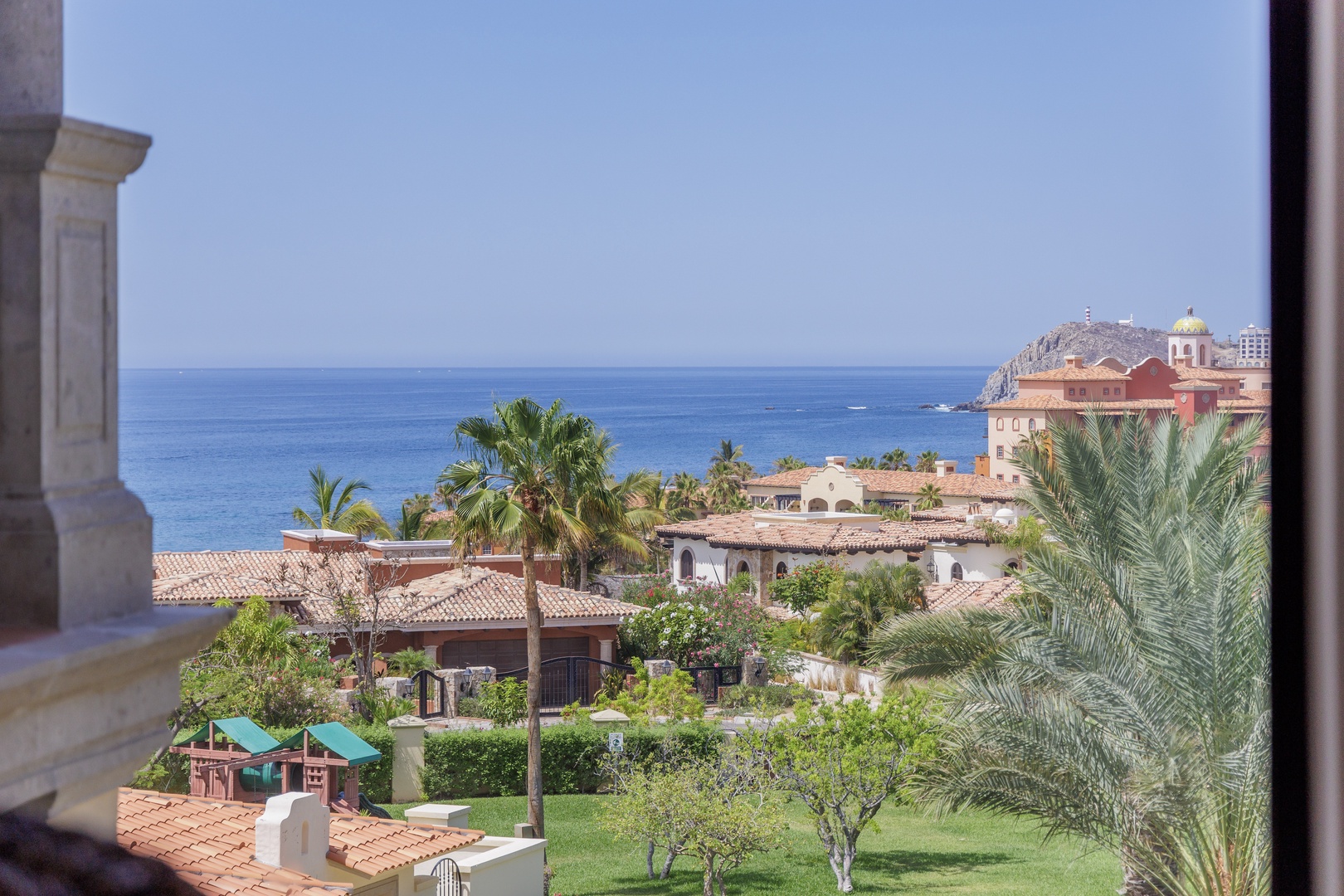 The property sits within a scenic coastal resort community, framed by lush gardens, palms, and Mediterranean-style architecture, with the Sea of Cortez horizon in the distance.